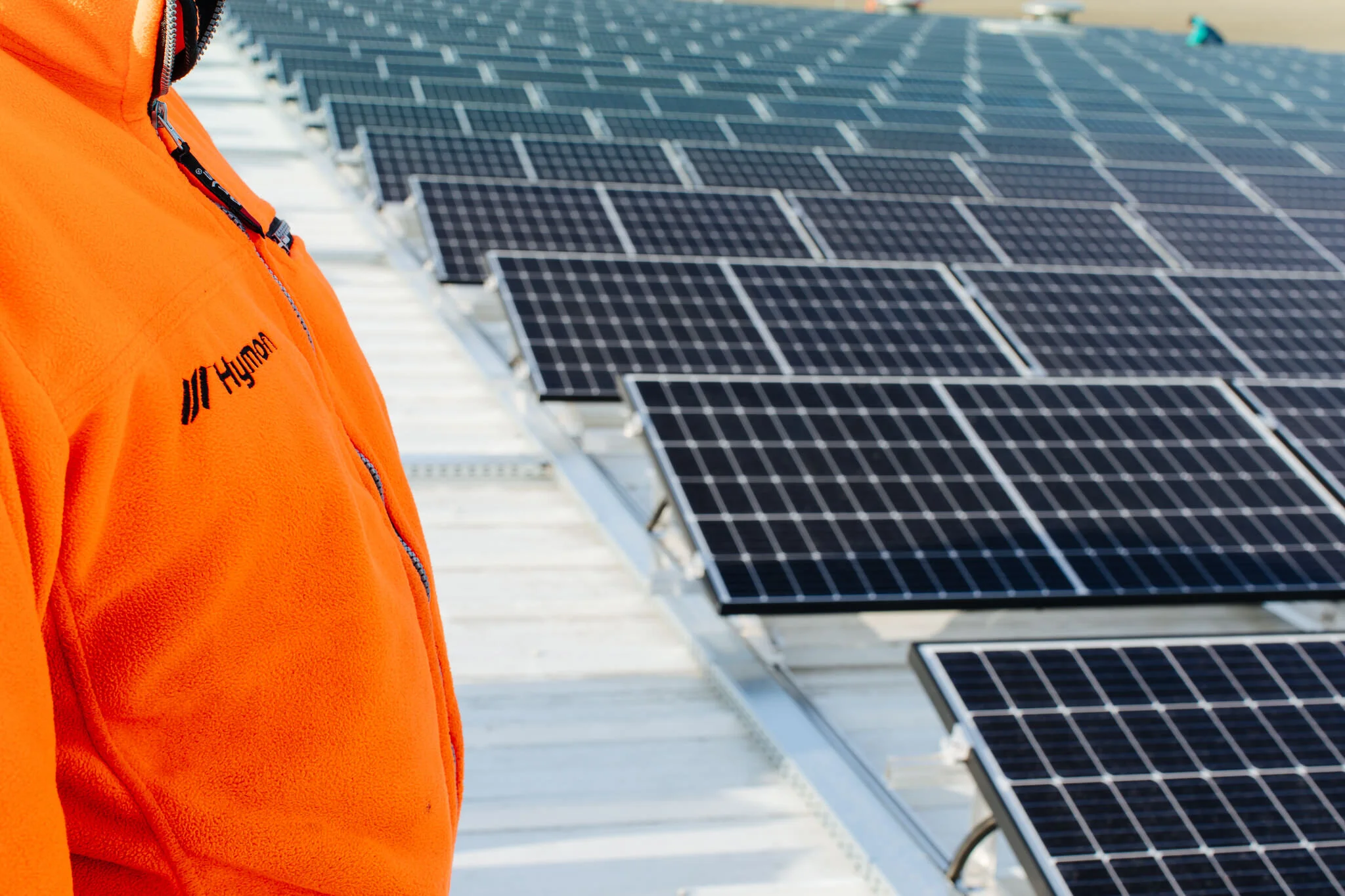 A person wearing an orange Hydron fleece jacket standing on a roof with black solar panels.