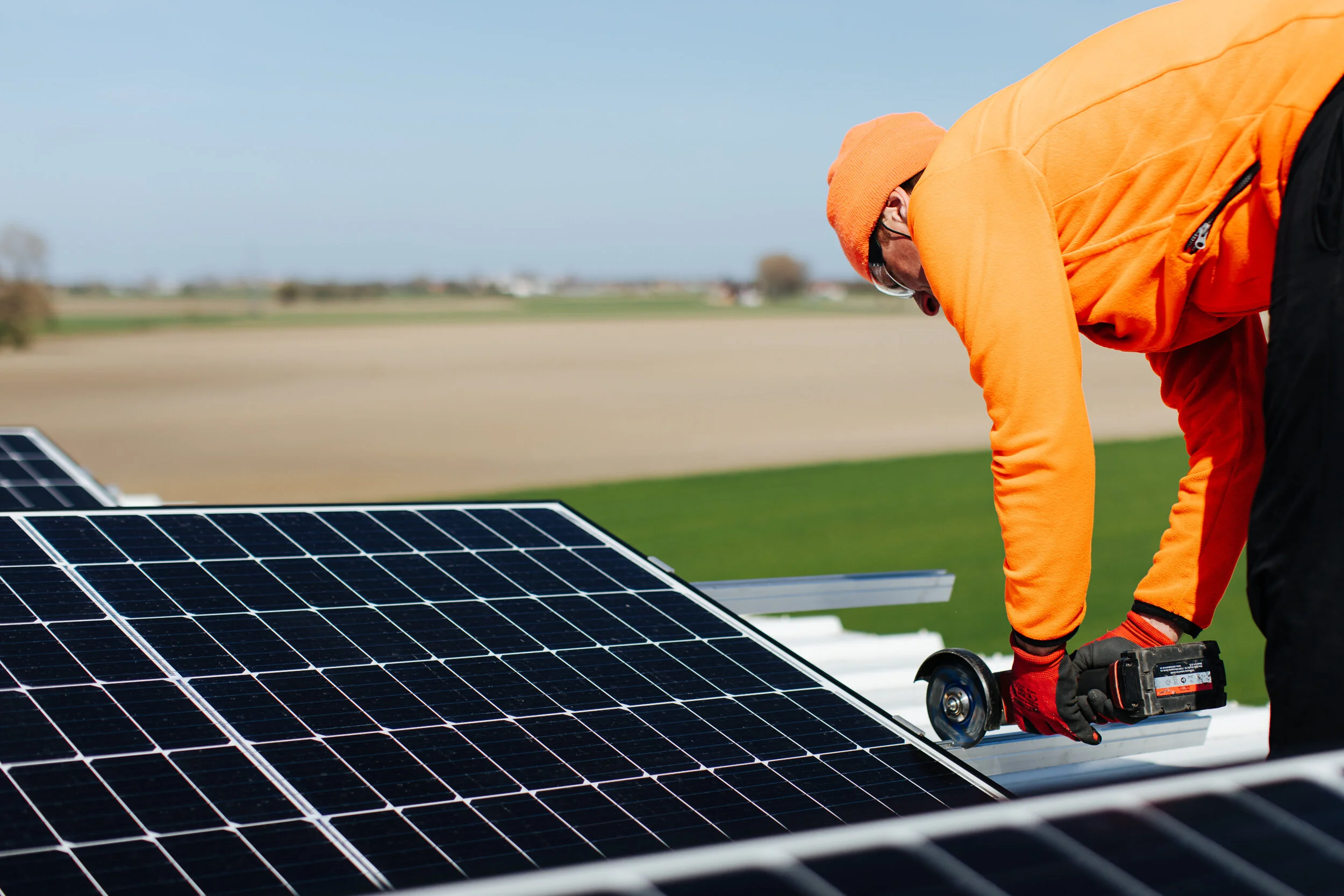 A man wearing an orange jacket, hat, and gloves is installing or maintaining solar panels outdoors on a sunny day in a rural area.