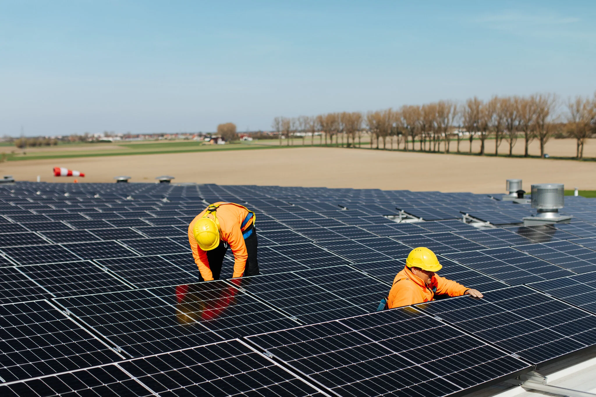 Two workers in orange uniforms and yellow helmets installing or maintaining solar panels on a large rooftop.