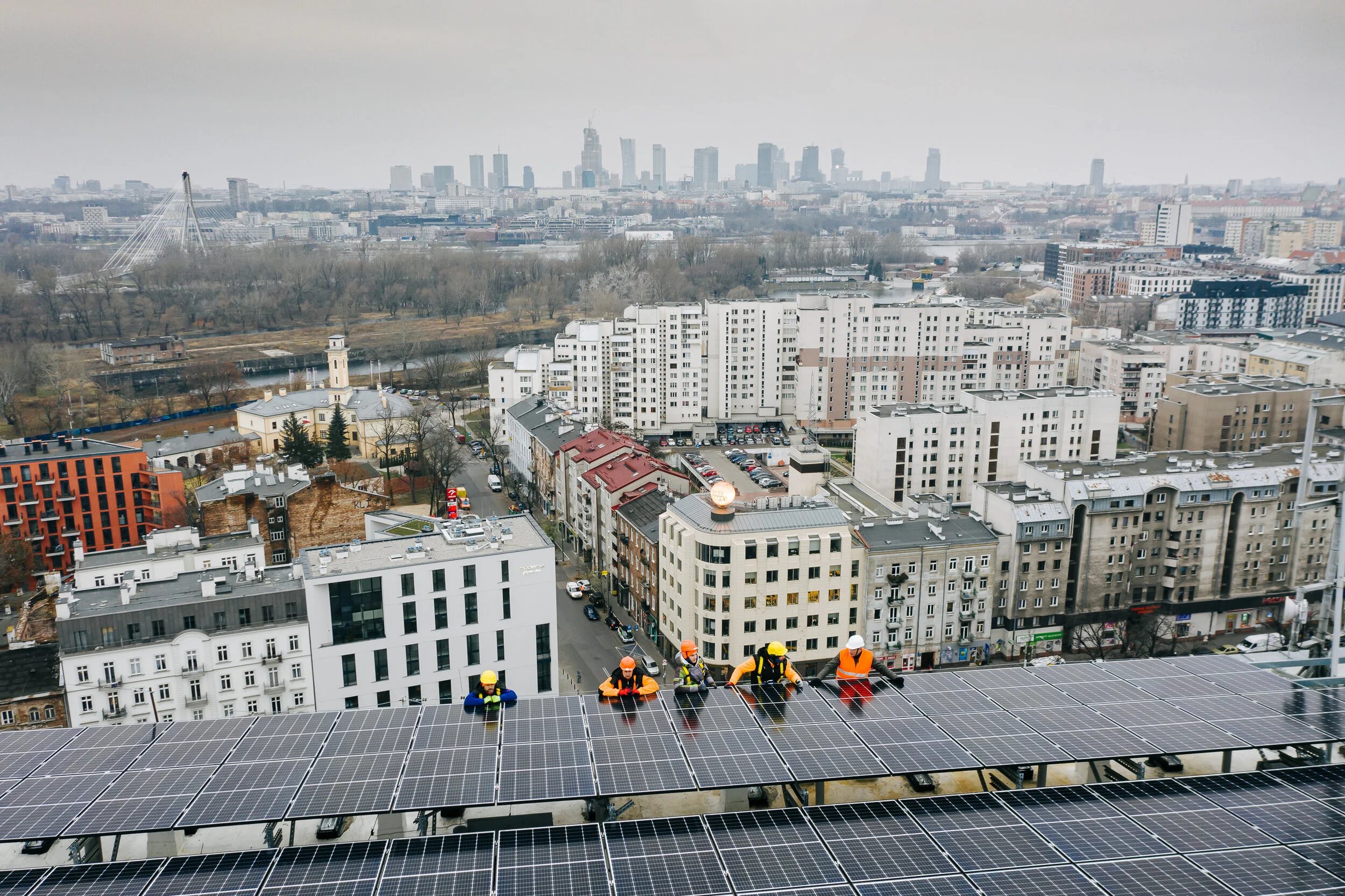 Construction workers wearing safety gear installing solar panels on a high-rise building roof with a city skyline in the background.
