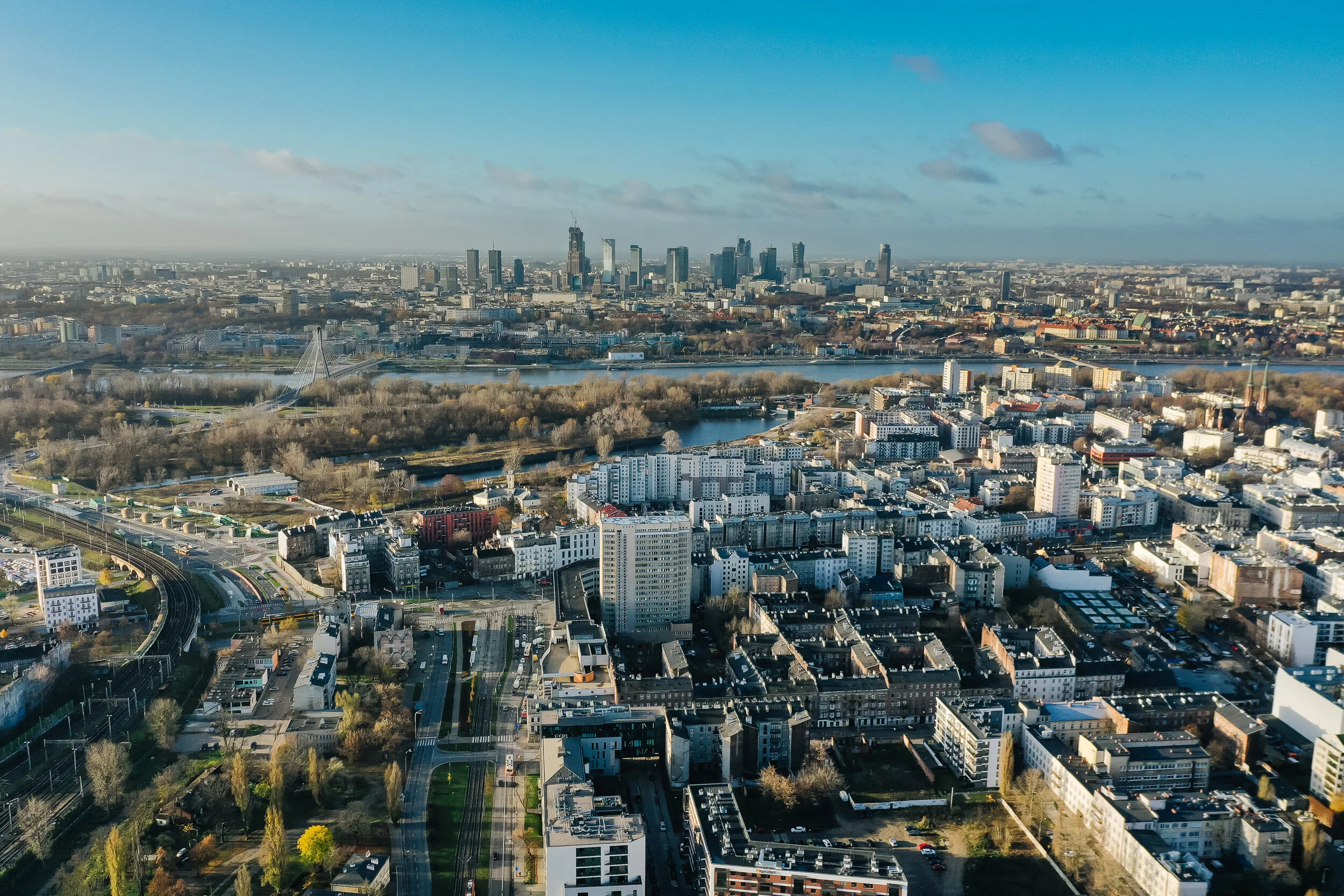 Aerial view of a cityscape with a river, numerous buildings, and a skyline with tall skyscrapers in the distance on a clear day.