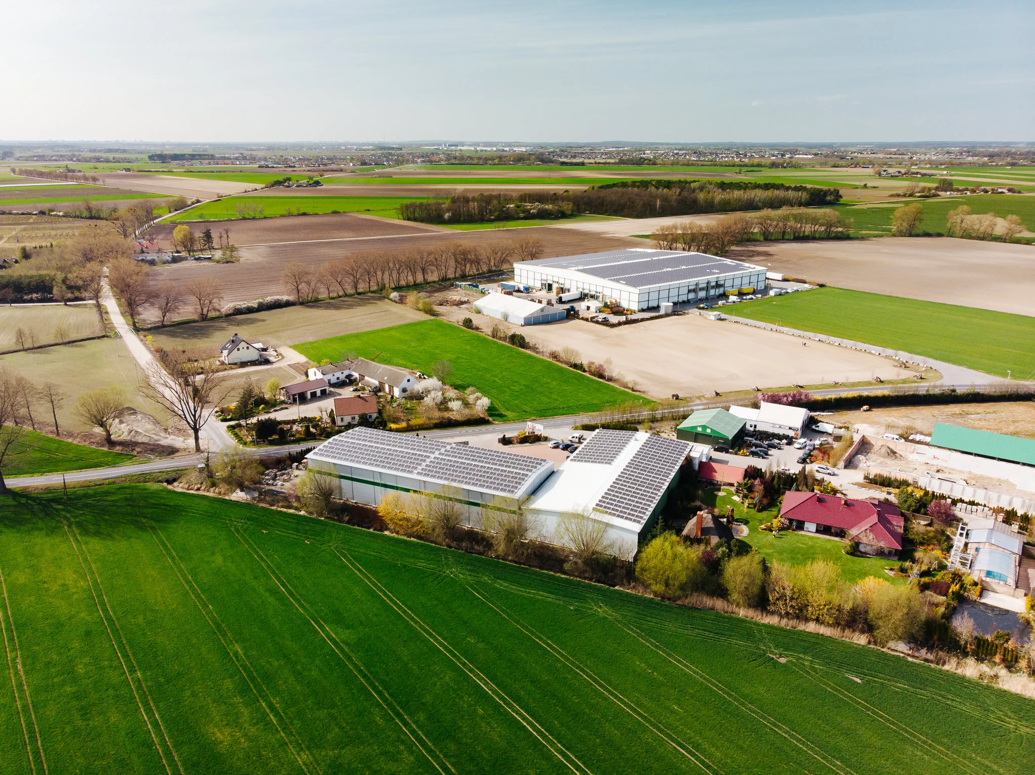 Aerial view of a rural area with farmhouses, green fields, and industrial buildings, some with solar panel rooftops, surrounded by trees and farmland.