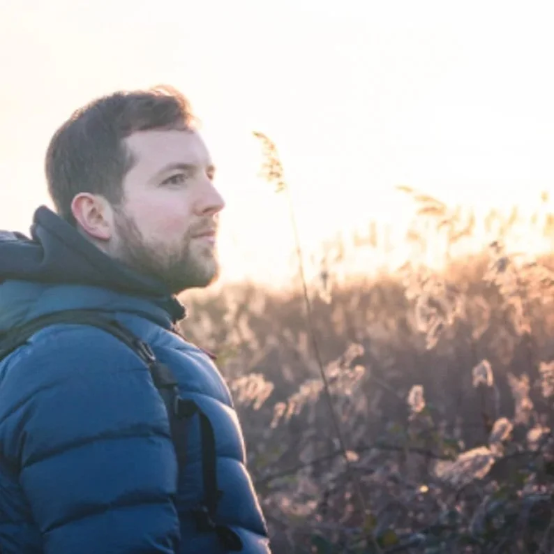 A man with a beard wearing a blue jacket and backpack looking to the left during sunset in a field with tall grasses.