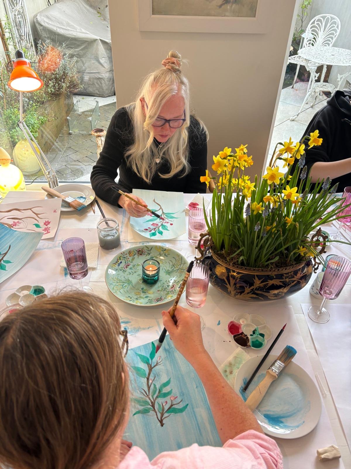 A group of elderly women sitting around a table involved in an art activity. Some are drawing on white sheets of paper, and glasses of champagne are on the table. There is a wooden sculpture in the center of the table, a large monitor screen on the wall, and a window with blinds letting in natural light.