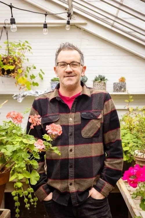 A man standing in a greenhouse or garden with pink and green plants around him, wearing glasses, a red shirt, and a brown and black plaid jacket, smiling with hands in pockets.
