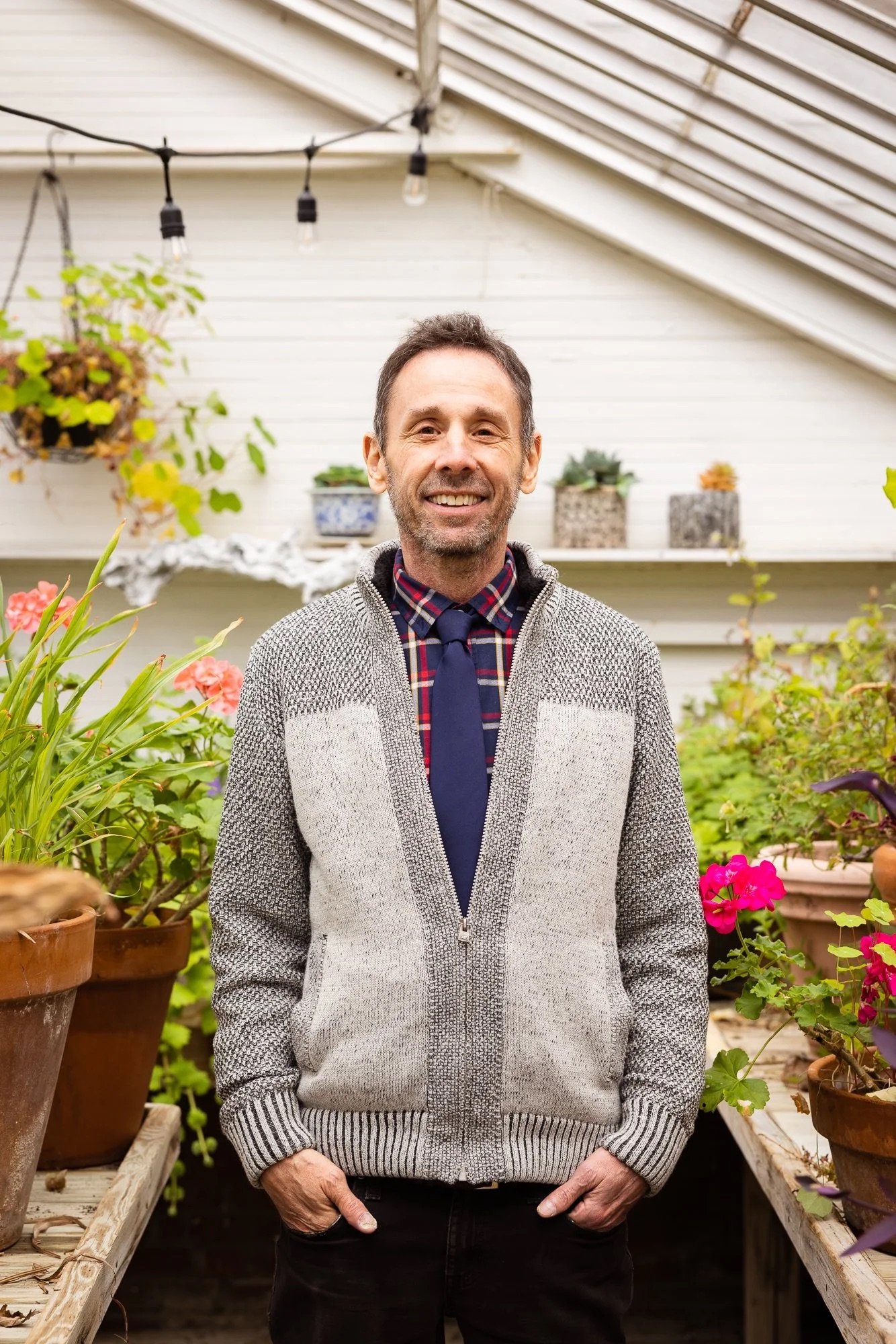 A smiling man standing in a greenhouse with potted plants and flowers around him.