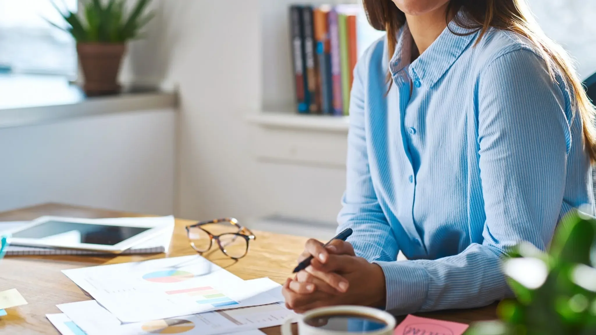 Frau im Büro sitzt an einem Tisch mit Dokumenten, einer Brille, einer Tasse Kaffee, einem Tablet und einem Laptop.