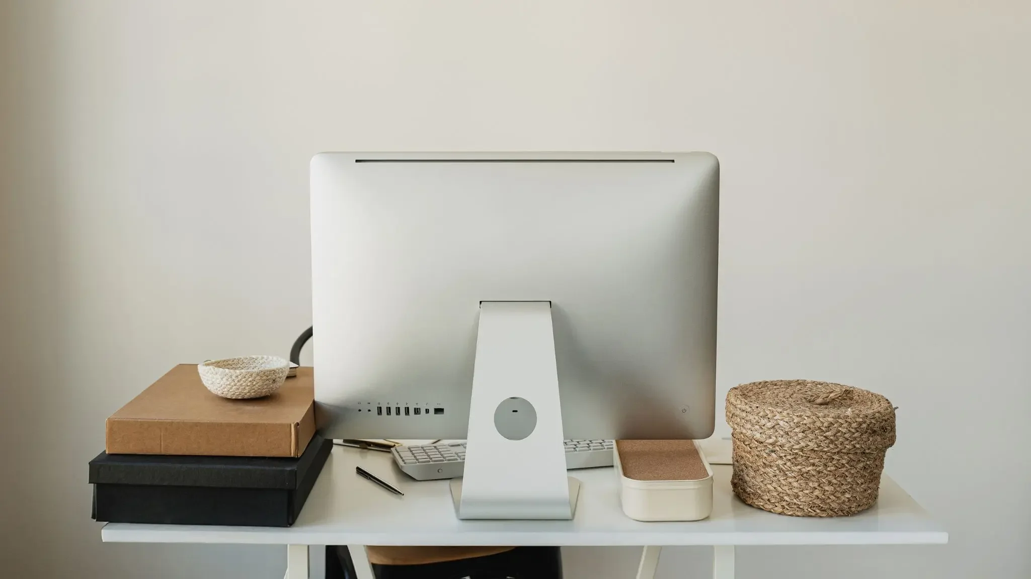 Schreibtisch mit einem iMac-Computer, drei gestapelten Boxen, einer Keramikkugel, einem weißen Tastatur, einem Stift, einem runden Lautsprecher, einem kleinen rechteckigen Korb und einer runden geflochtenen Aufbewahrungsbox.