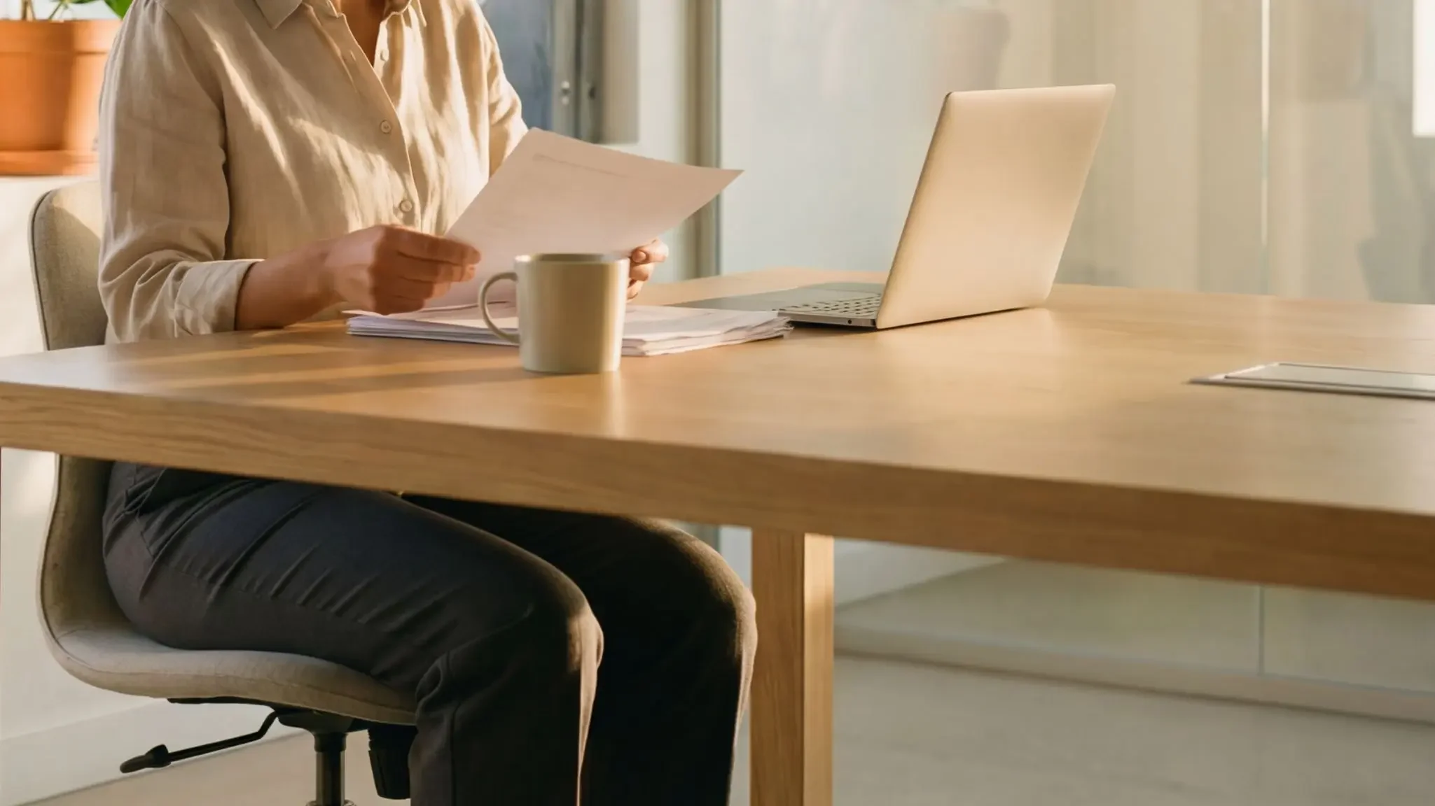 Frau sitzt an einem Holztisch, liest Dokumente, mit Laptop, Kaffeetasse und einem Notizbuch auf dem Tisch.