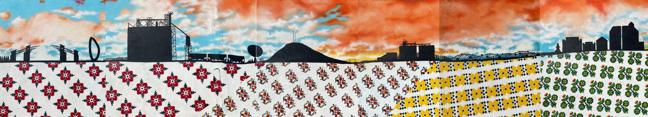 A city skyline with industrial buildings and a mountain in the background, above a decorative fence with colorful floral tiles, against a partly cloudy sky at sunset.