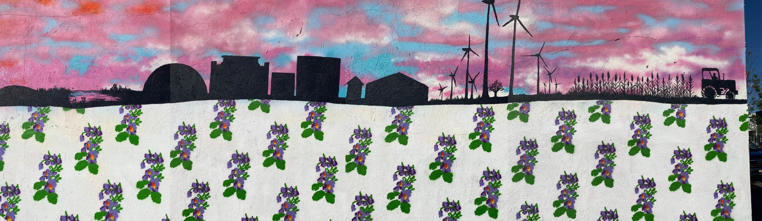A mural depicting a city skyline with wind turbines and a tractor against a pink and blue sky, with a foreground of purple and green flowers painted on a white wall.