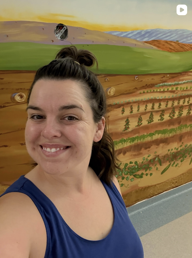 Artist Carly Gordon taking a selfie in front of a colorful mural depicting a landscape of fields, hills, and a sunset.