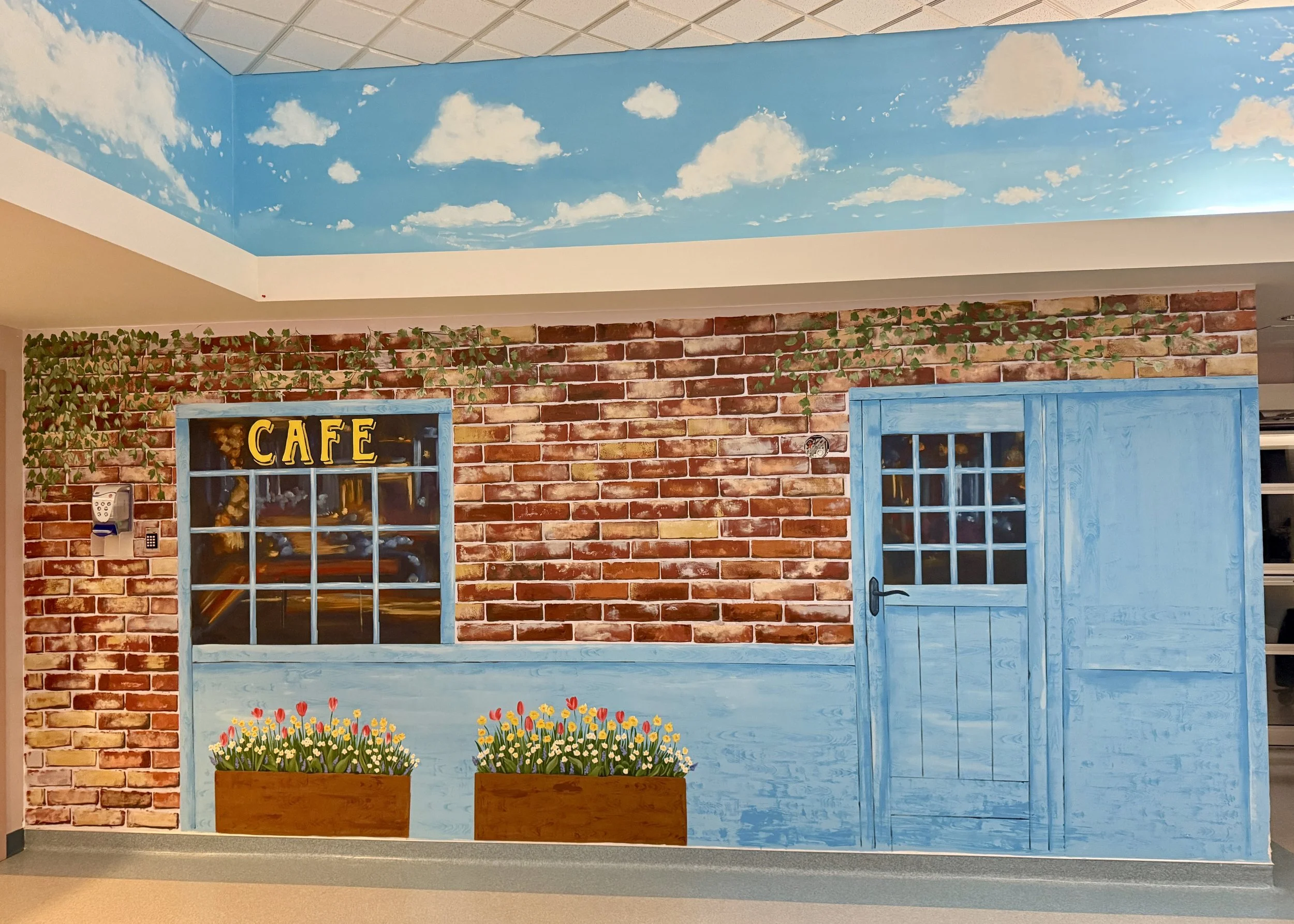 Wall painted mural of a brick building with a blue door, a window with the word 'Café' above it, flower boxes with tulips, and a sky ceiling with white clouds.