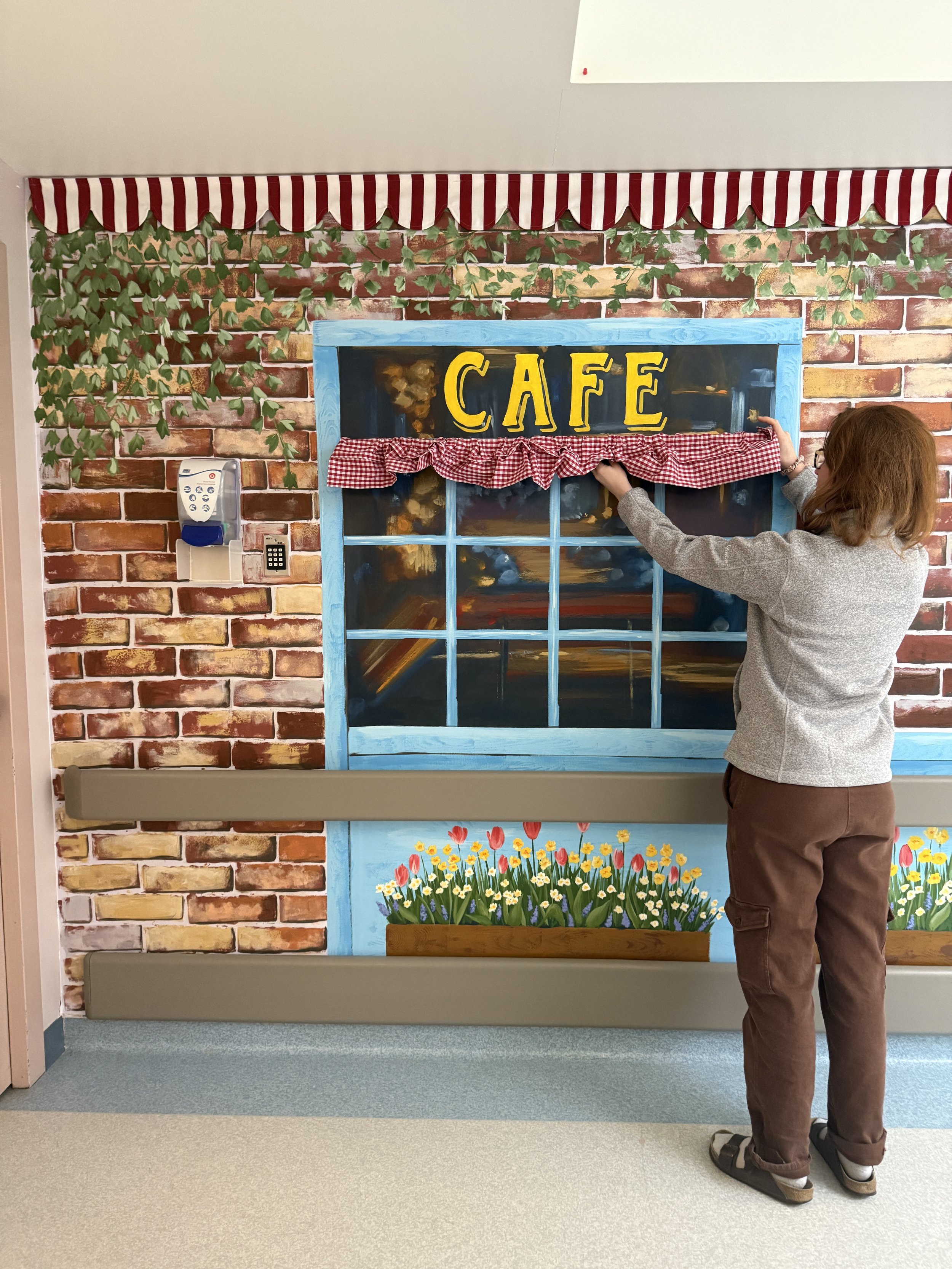A person is adjusting a painted window with the word 'CAFE' on it in front of a brick wall. The wall has painted green ivy and a flower bed below the window. There is a red and white checkered curtain hanging above the window, and a red and white striped awning at the top.