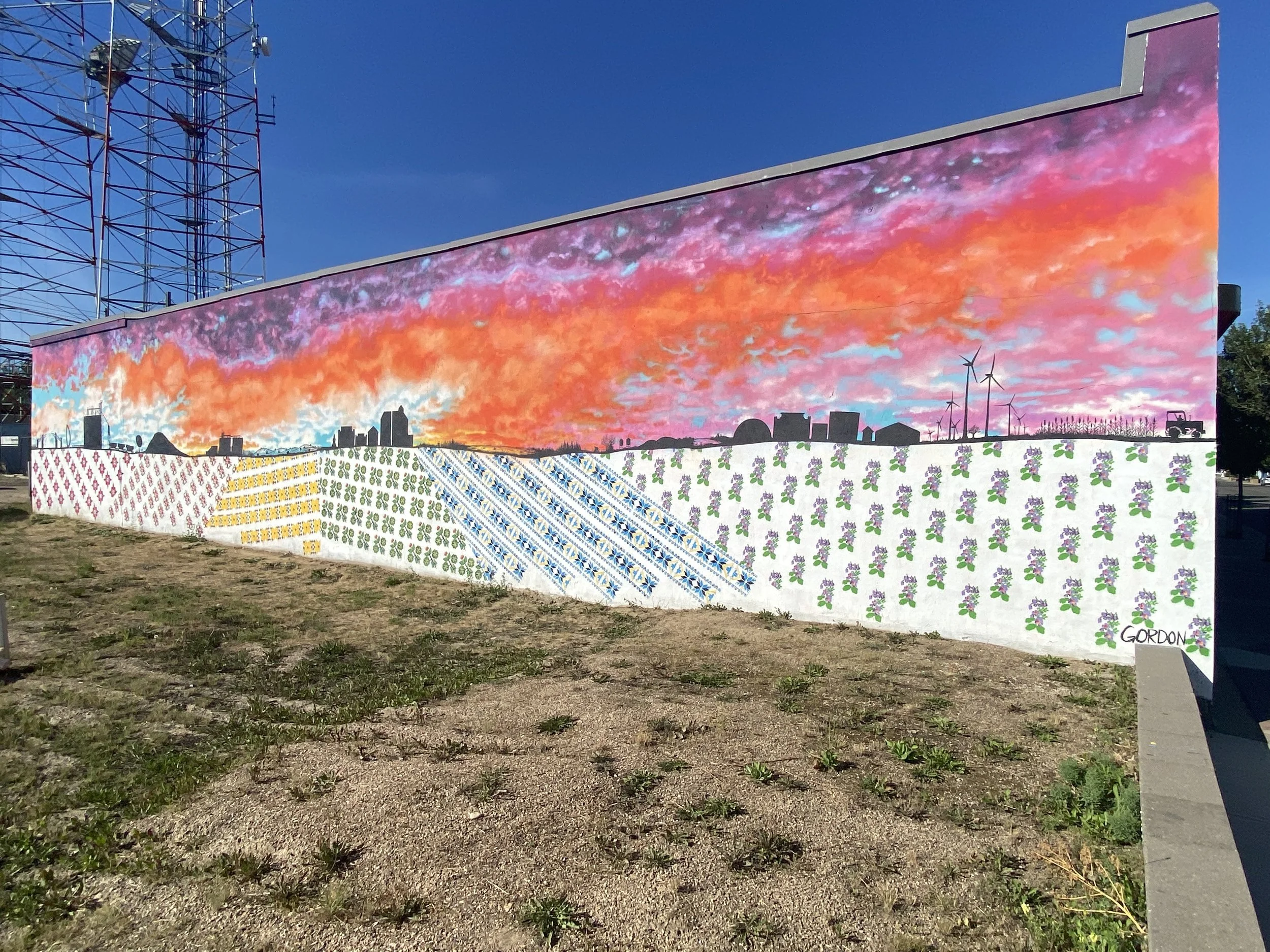 Large outdoor mural with a city skyline silhouette against a colorful sunset sky, decorated with wind turbines, and patterns of flowers on the lower part of the wall.