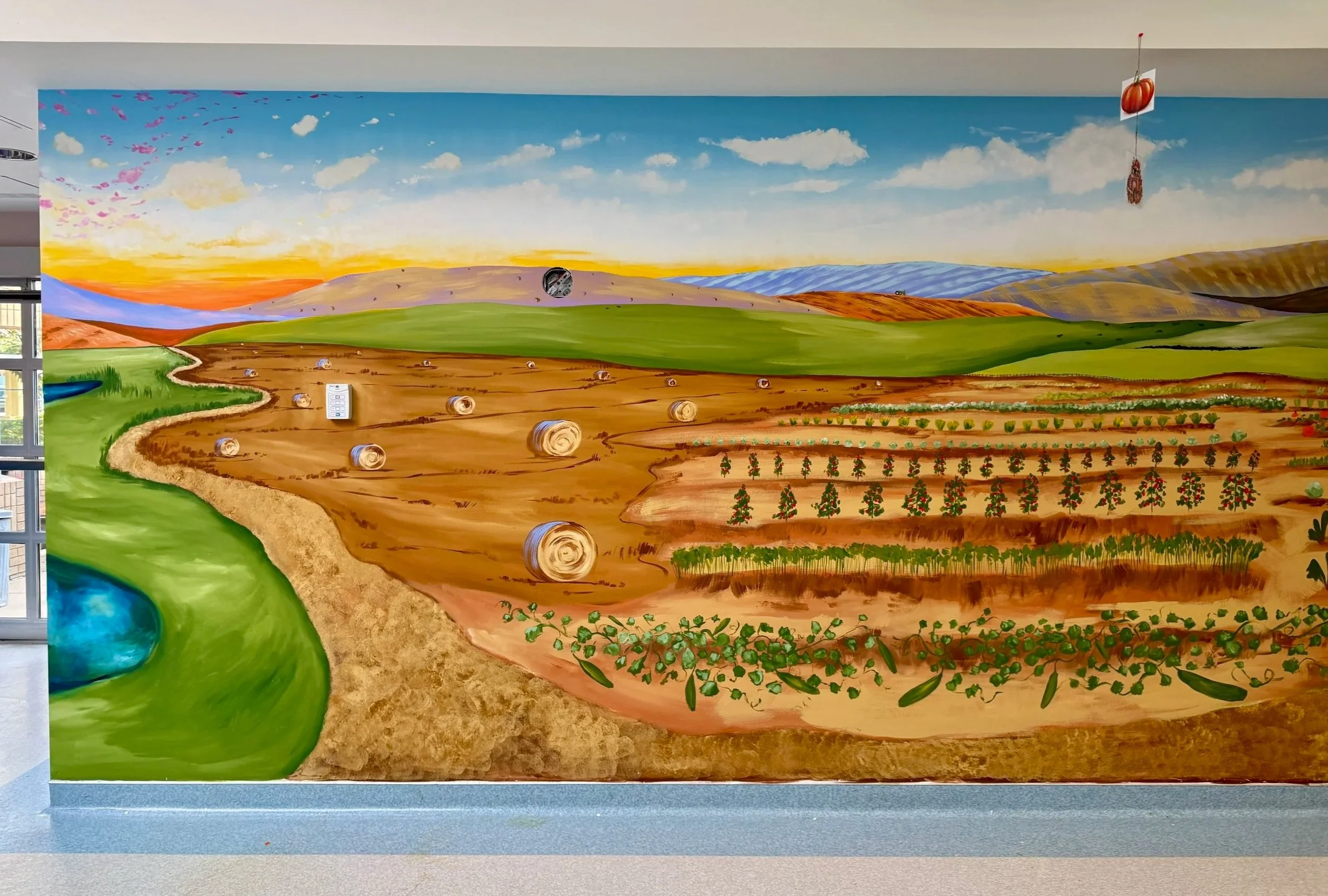 Colorful mural depicting a farm scene with hay bales, cultivated fields, green hills, and a blue sky with clouds and sunset.