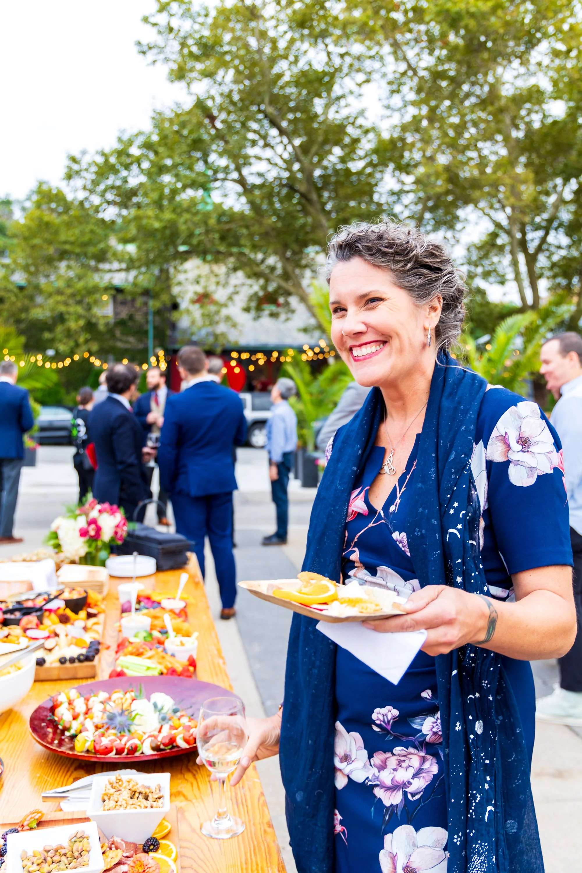 A woman smiling and holding a plate of food at an outdoor gathering with a table full of desserts and a group of people in the background.