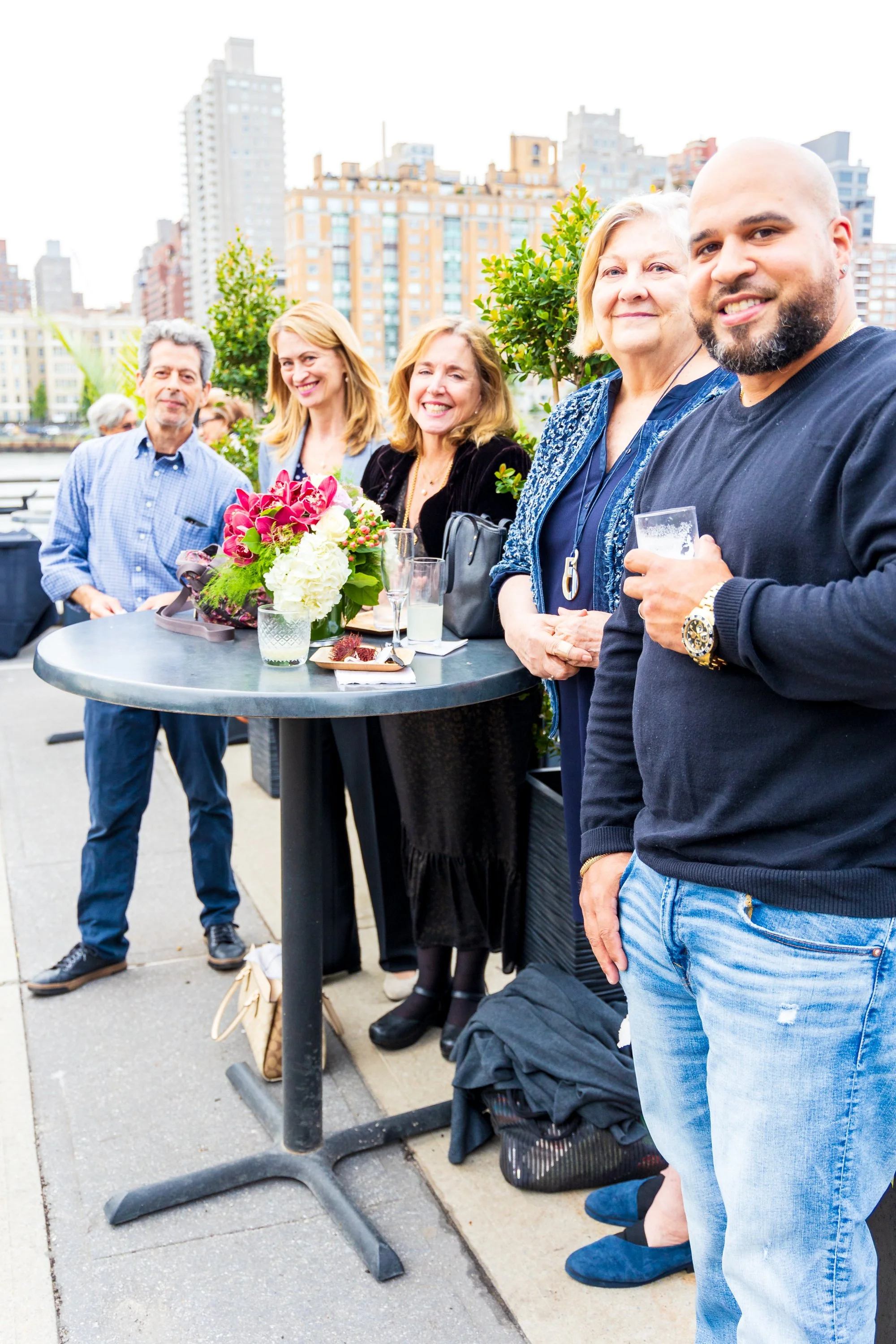 A group of six people gathered around a high-top table outdoors, with city buildings in the background. The table has a bouquet of pink and white flowers. Everyone is smiling and enjoying drinks, celebrating a social occasion.