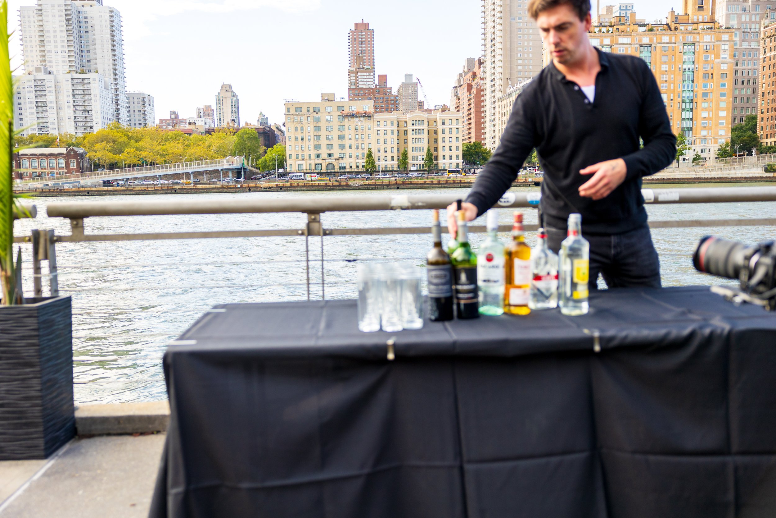 A man arranging bottles of alcohol and glasses on a black table with a city skyline and river in the background.
