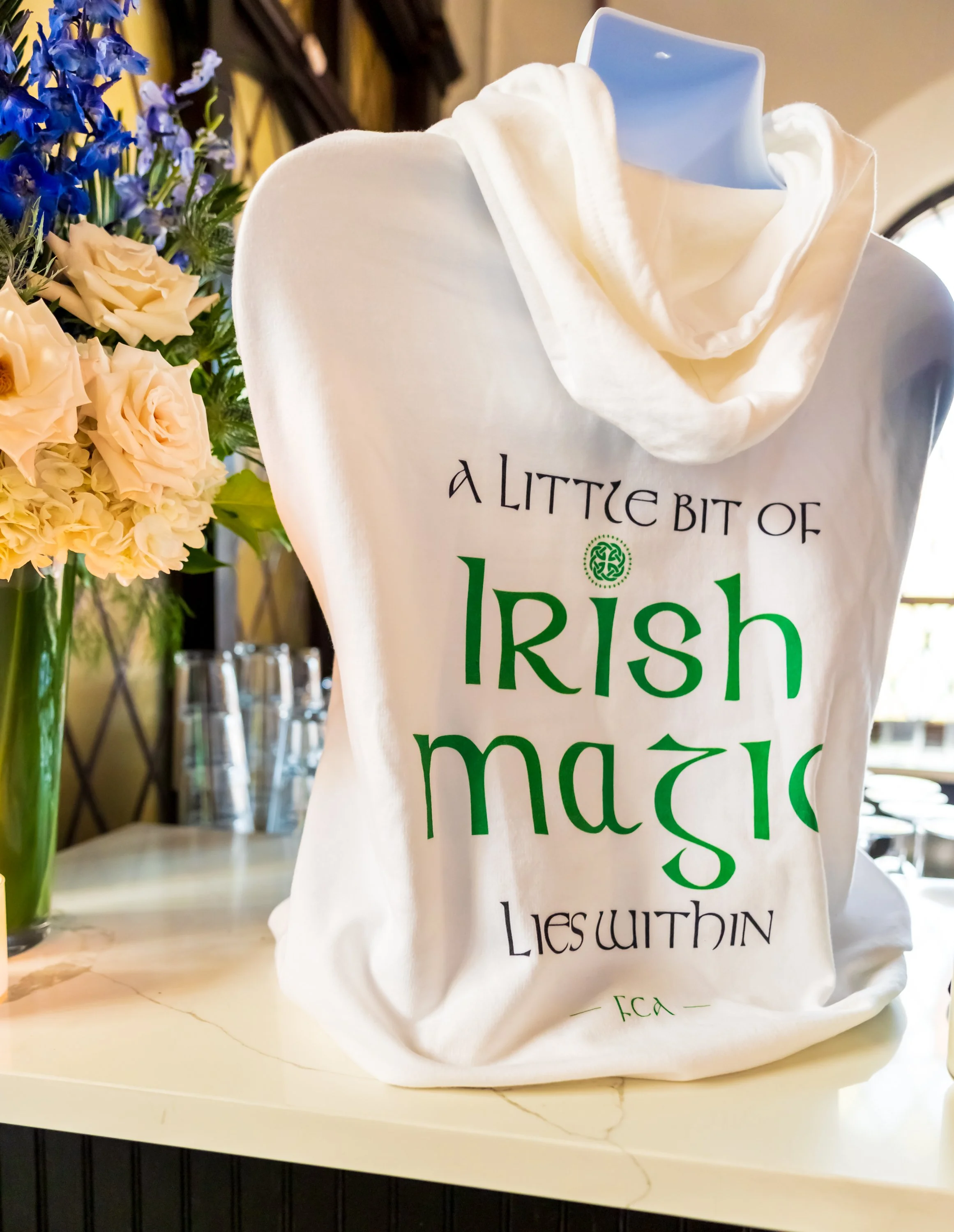 White tote bag with green and black text that reads 'A LITTLE BIT OF IRISH magic LIES WITHIN' displayed on a white marble countertop, with a bouquet of pink roses and purple flowers in a glass vase nearby.