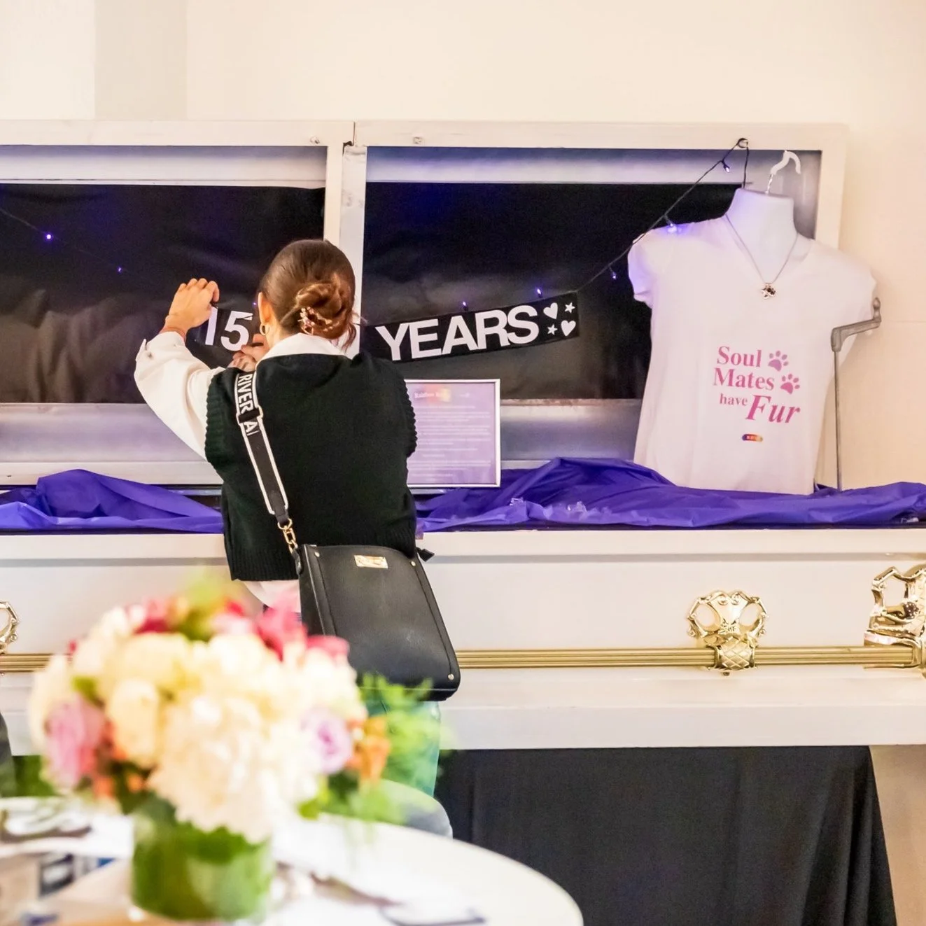 Person placing a sign that reads "15 YEARS" inside a display case, with a white t-shirt on a hanger that says "Soul Mates have Fur" and a bouquet of flowers in the foreground.