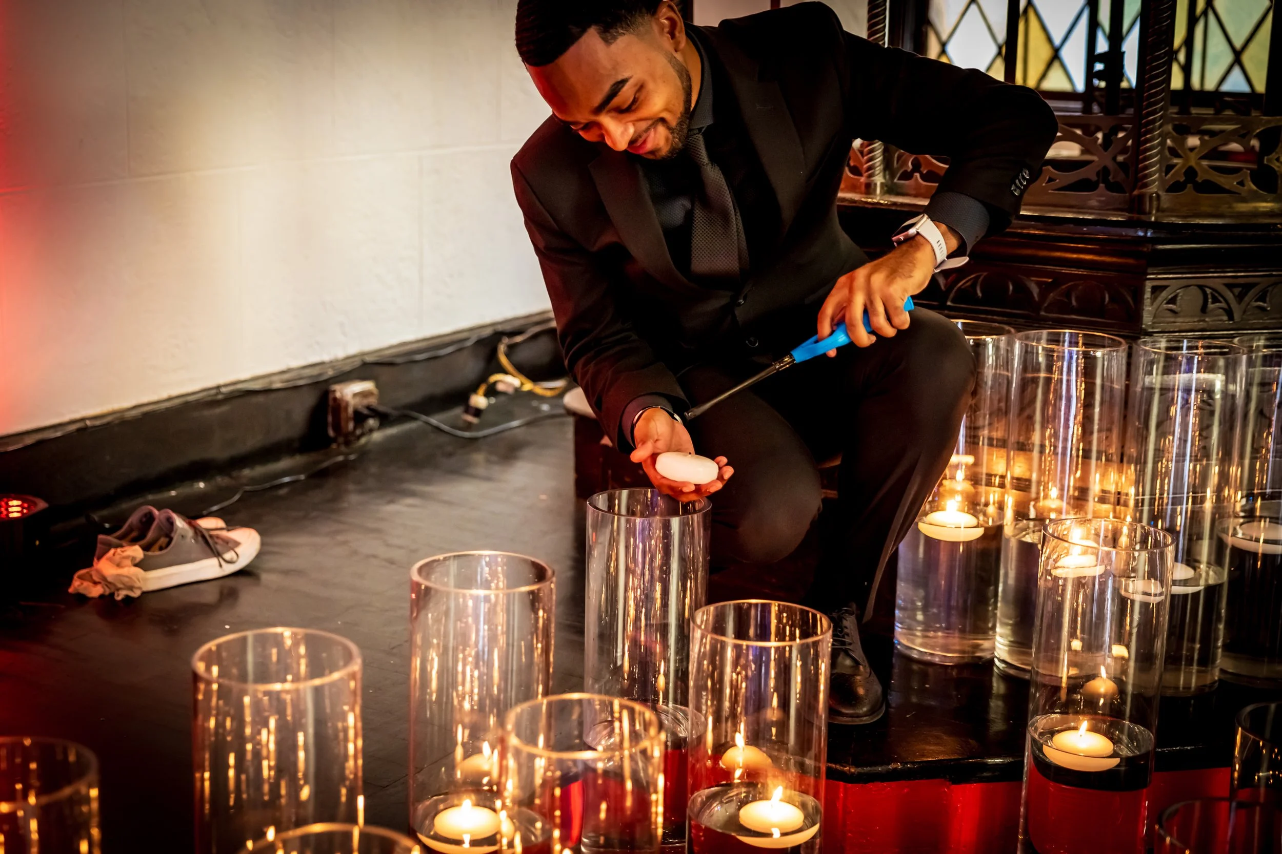 A man in a black suit and tie crouches on the floor, lighting candles in glass holders using a long lighter. The setting appears to be indoors with a decorative wooden railing in the background and candles arranged on the floor, creating a warm ambia