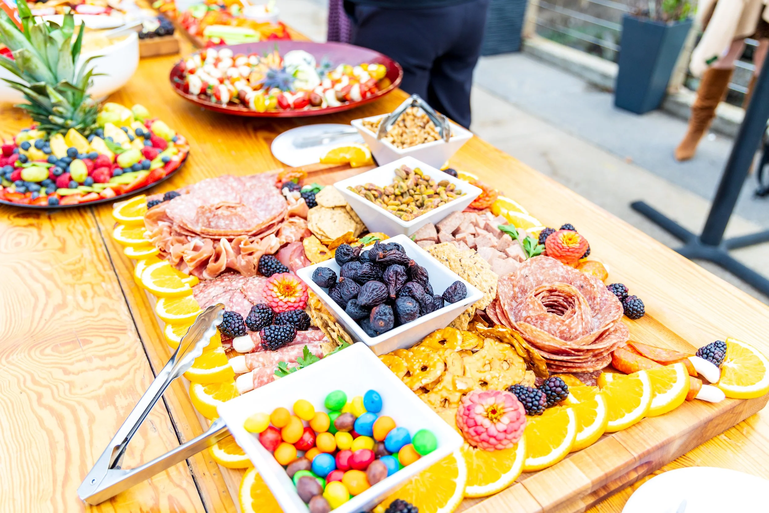 A wooden table with assorted fruits, candies, nuts, and cured meats arranged for a buffet or gathering, outdoors.