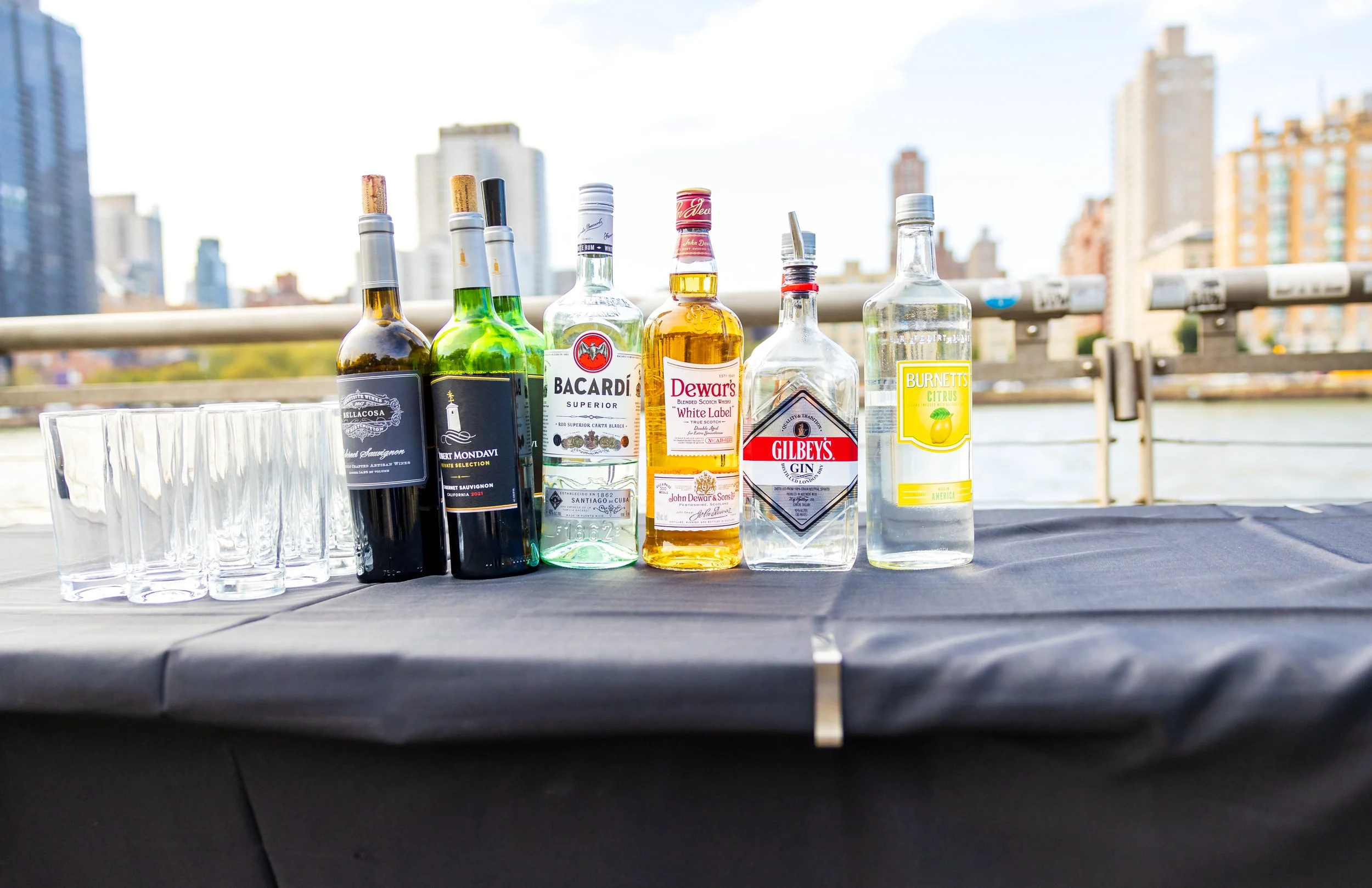 Selection of liquor bottles including Bacardi, Dewar's, Gilbey's, and Burnett's, along with empty shot glasses, on a black table outdoors with a city skyline in the background.
