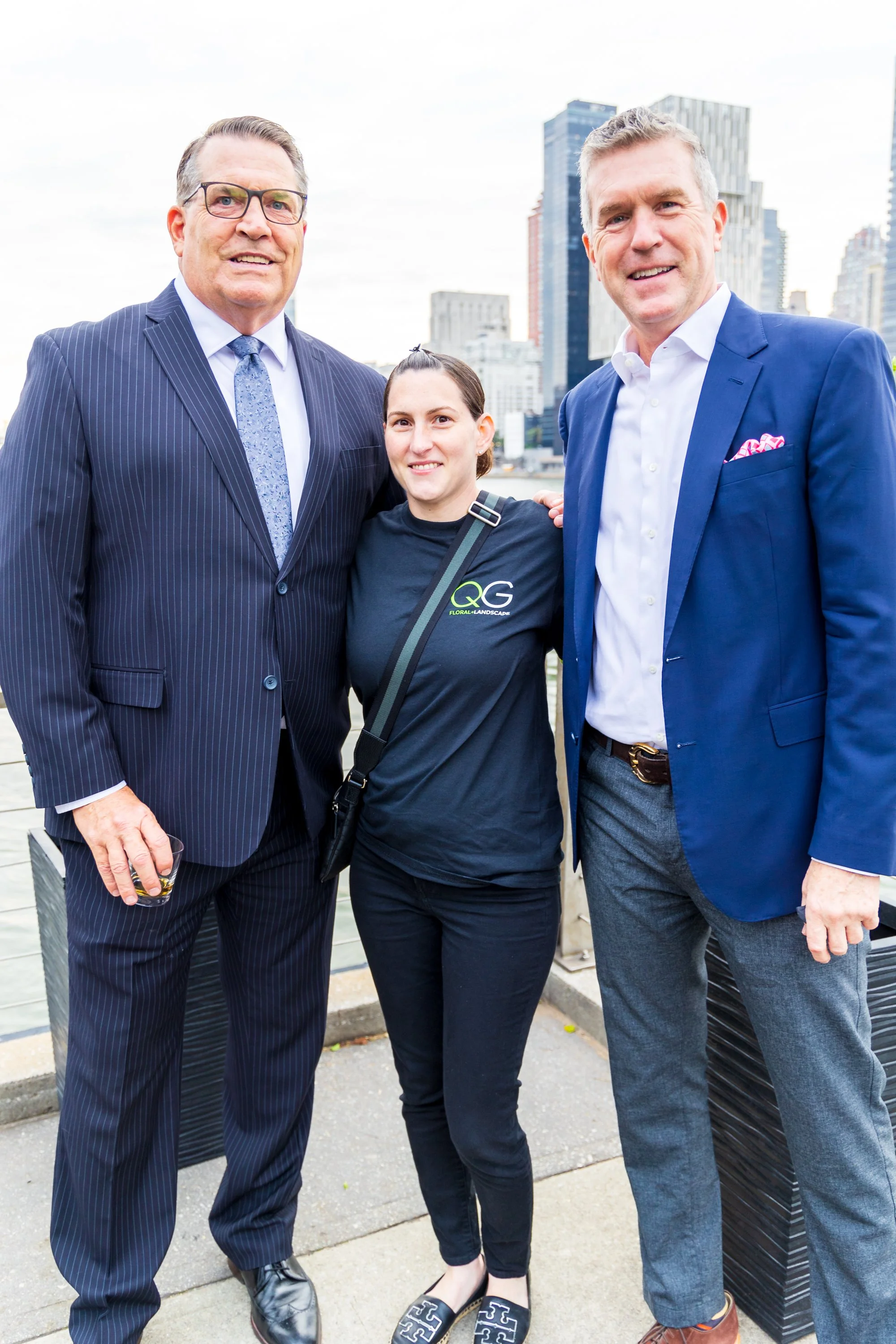Three people smiling outdoors with cityscape background, two men in suits and a woman in casual attire.