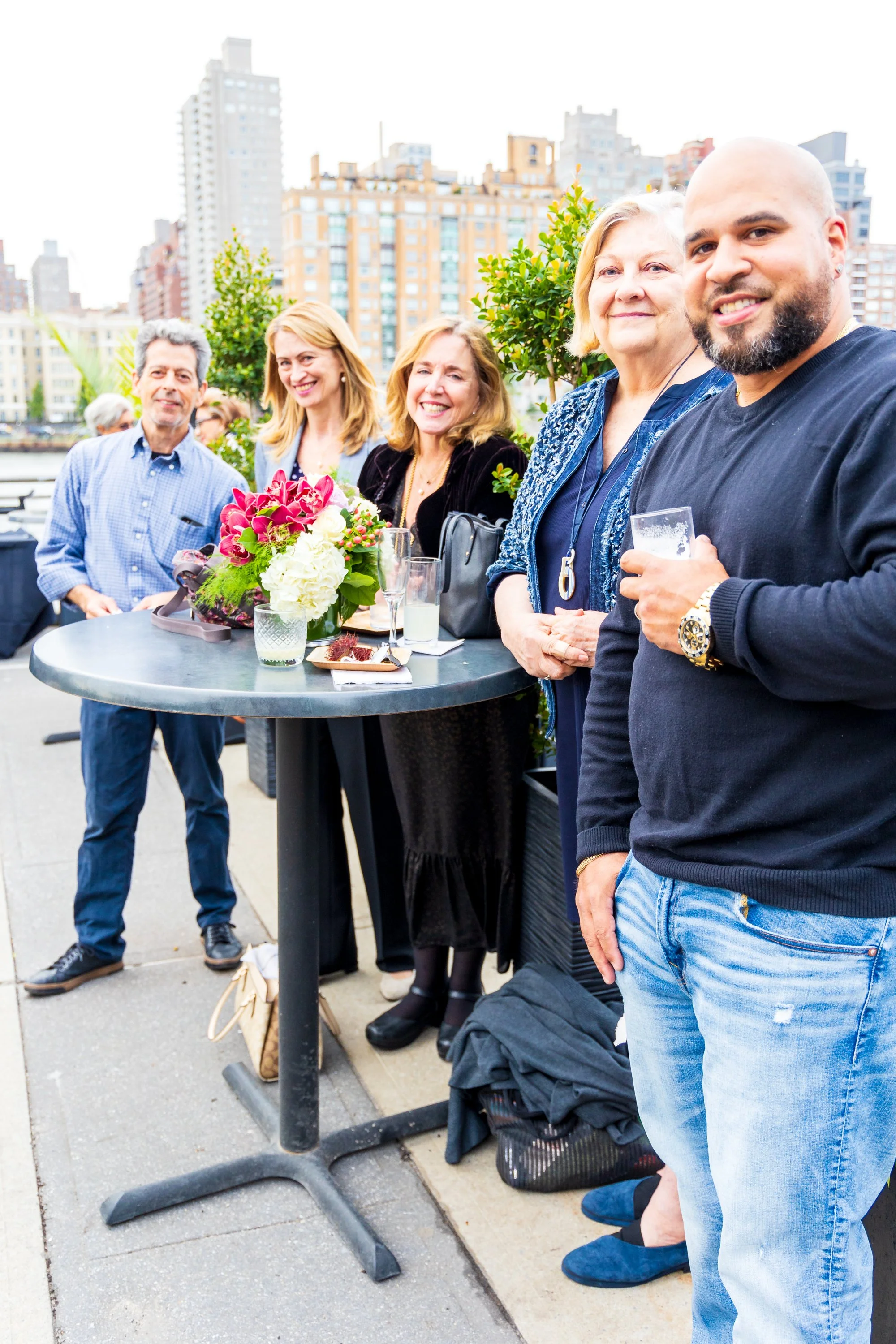 Group of six people standing around a high-top table outdoors, smiling, with city buildings in the background, and a floral centerpiece on the table.
