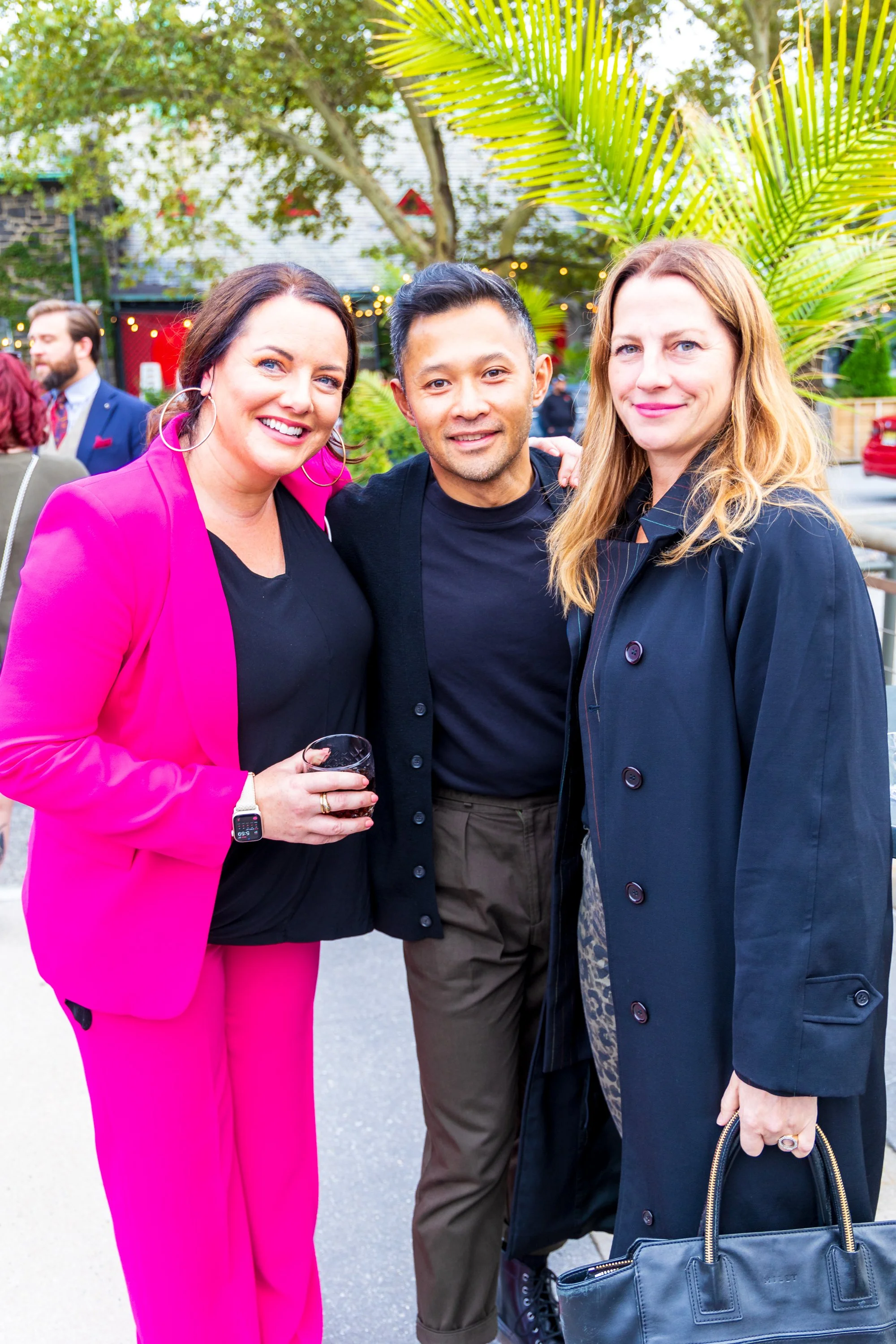 Three people smiling outdoors during an event, with one woman holding a glass of wine, a man in the middle, and a woman on the right holding a handbag, with trees and string lights in the background.