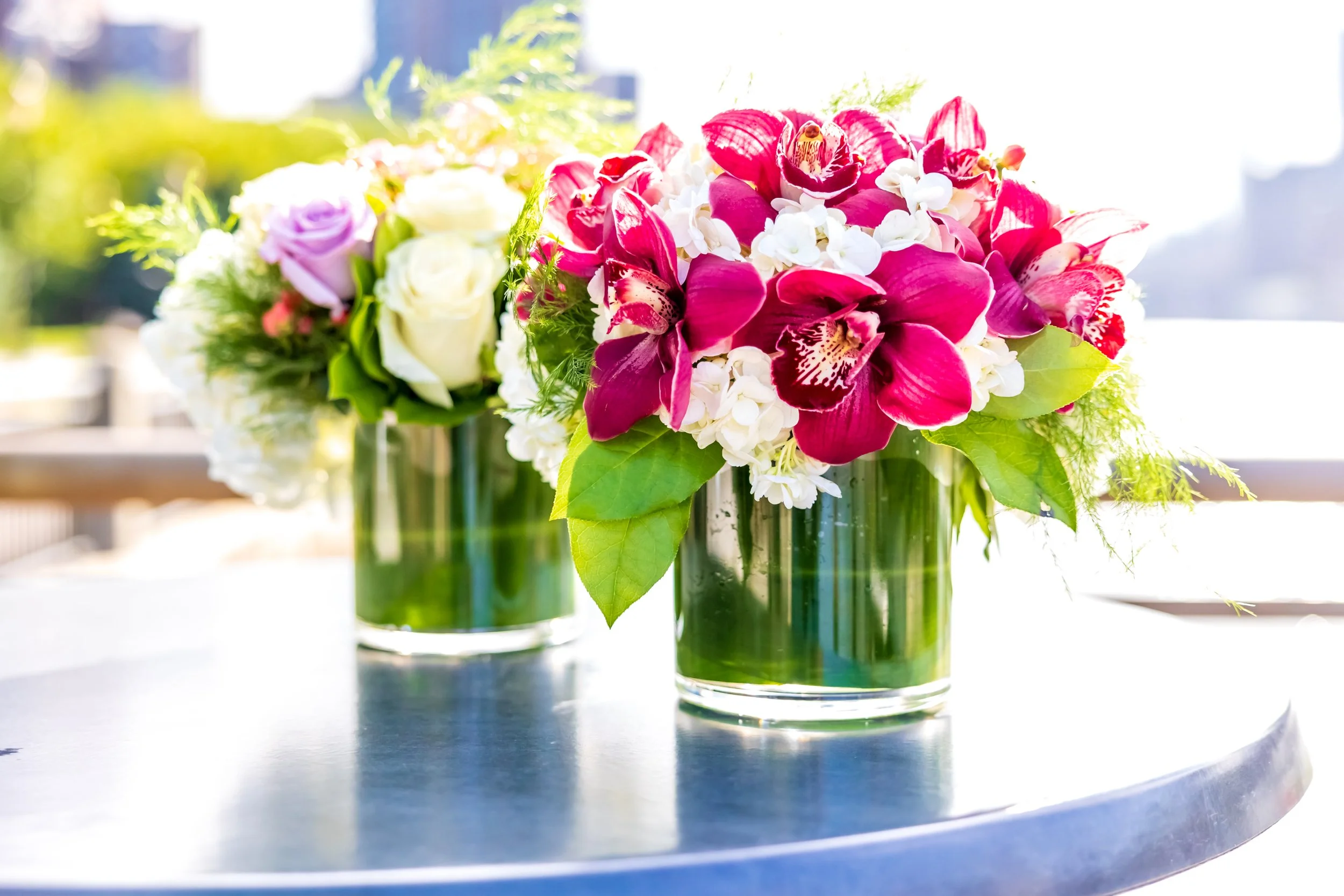 Two glass vases with colorful flower arrangements, including pink orchids, white roses, and purple and white flowers on a table outdoors.