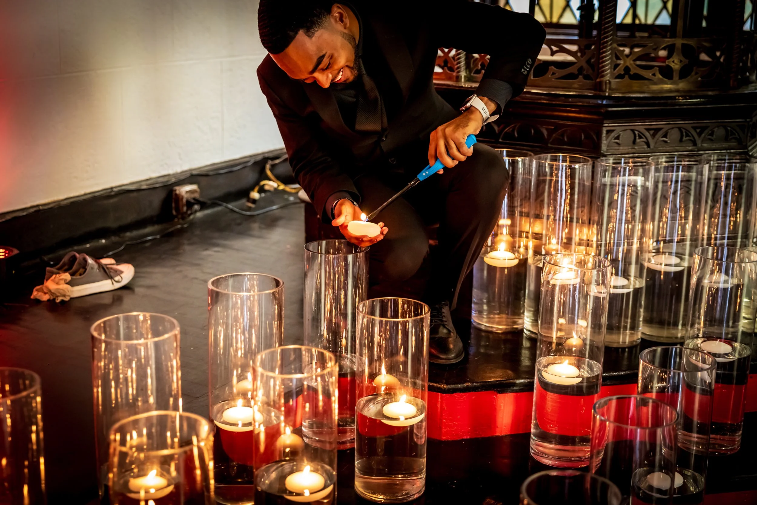 Man in a black suit lighting a candle surrounded by glass candle holders at an indoor event.