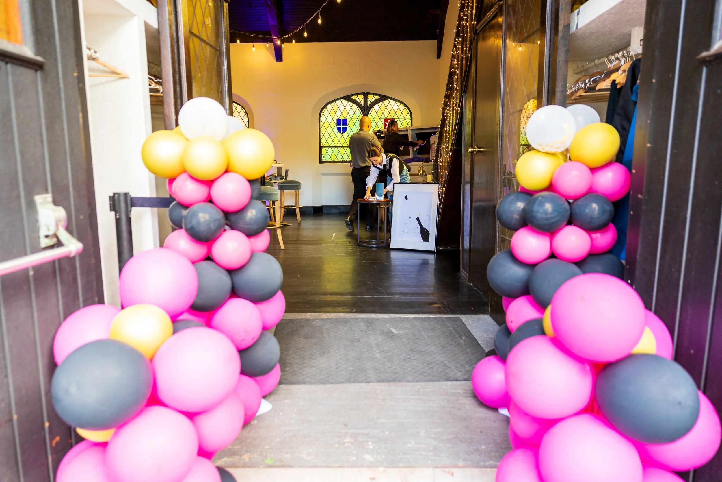 Decorative balloon arch in pink, black, yellow, and white at the entrance of a cozy event space with people preparing inside.