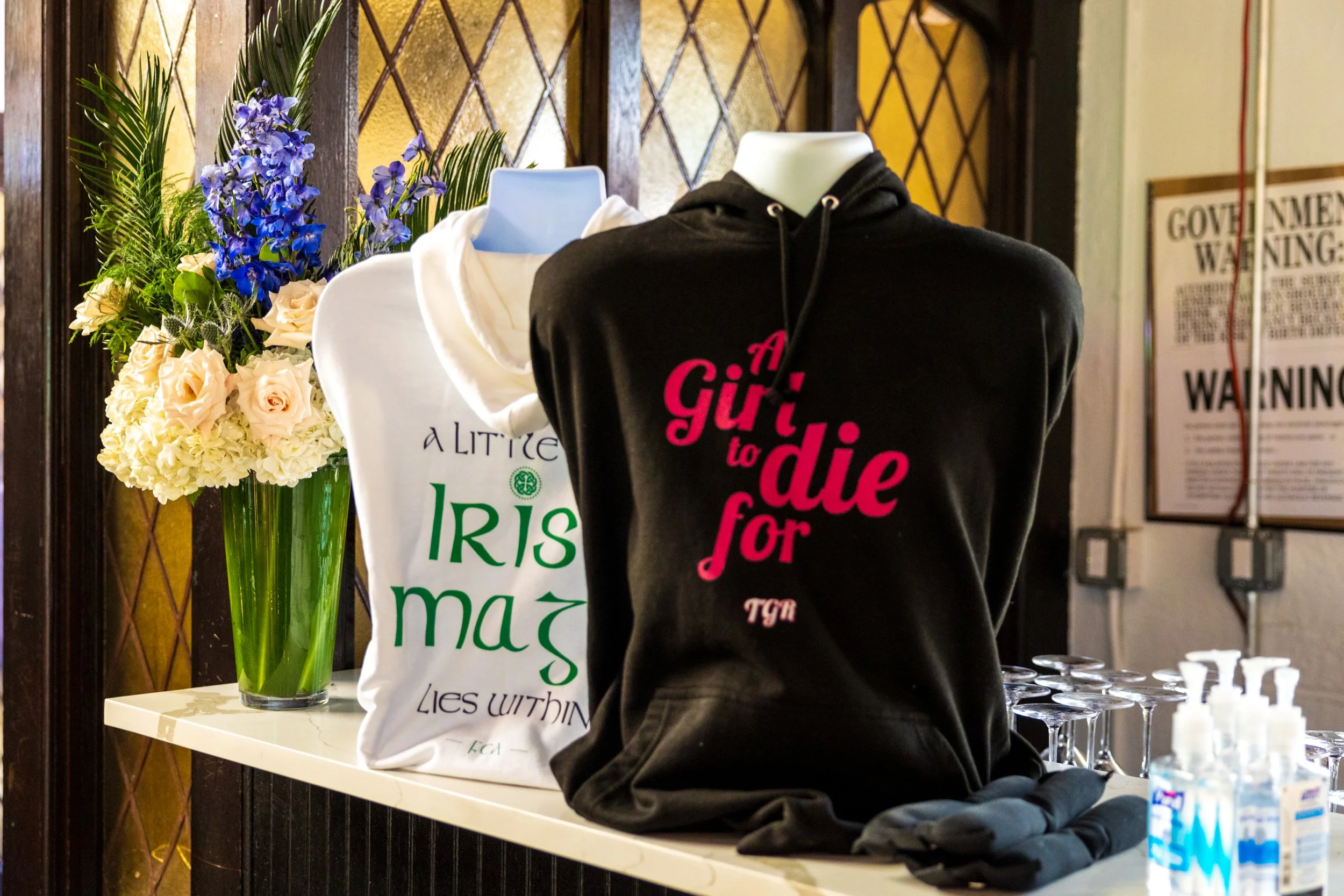 Black hoodie with pink text reading "A Girl to Die for" hanging on a mannequin next to a white hoodie with green and black text and a floral tote bag with the words "A Little Iris" on a table, with a bouquet of flowers and bottles of hand sanitizer n