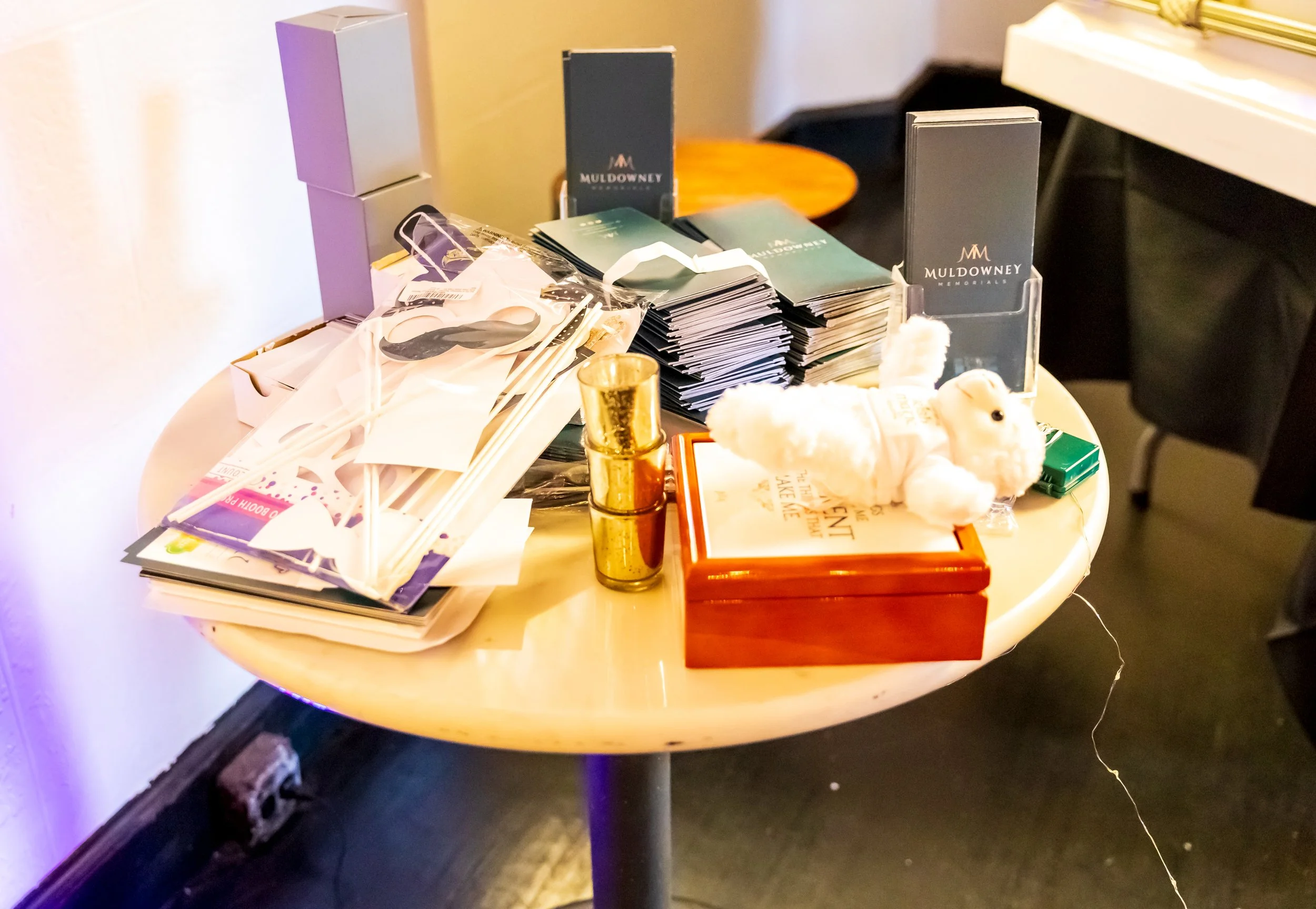 Round table with assorted promotional items including drink stirrers, brochures, a plush teddy bear, and branded boxes from Mul Downey Jewelers.