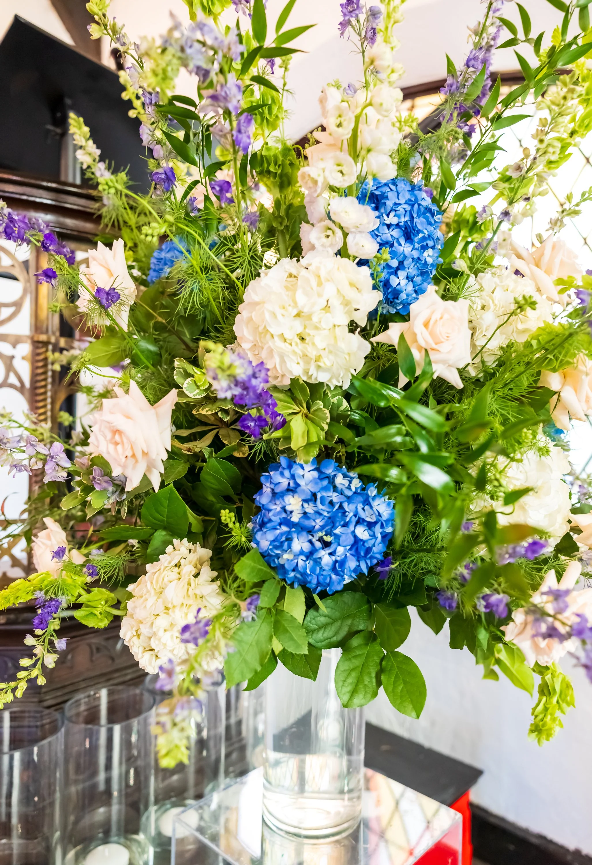 A colorful bouquet of flowers in a clear glass vase, containing blue hydrangeas, white hydrangeas, peach roses, purple delphiniums, and various greenery.