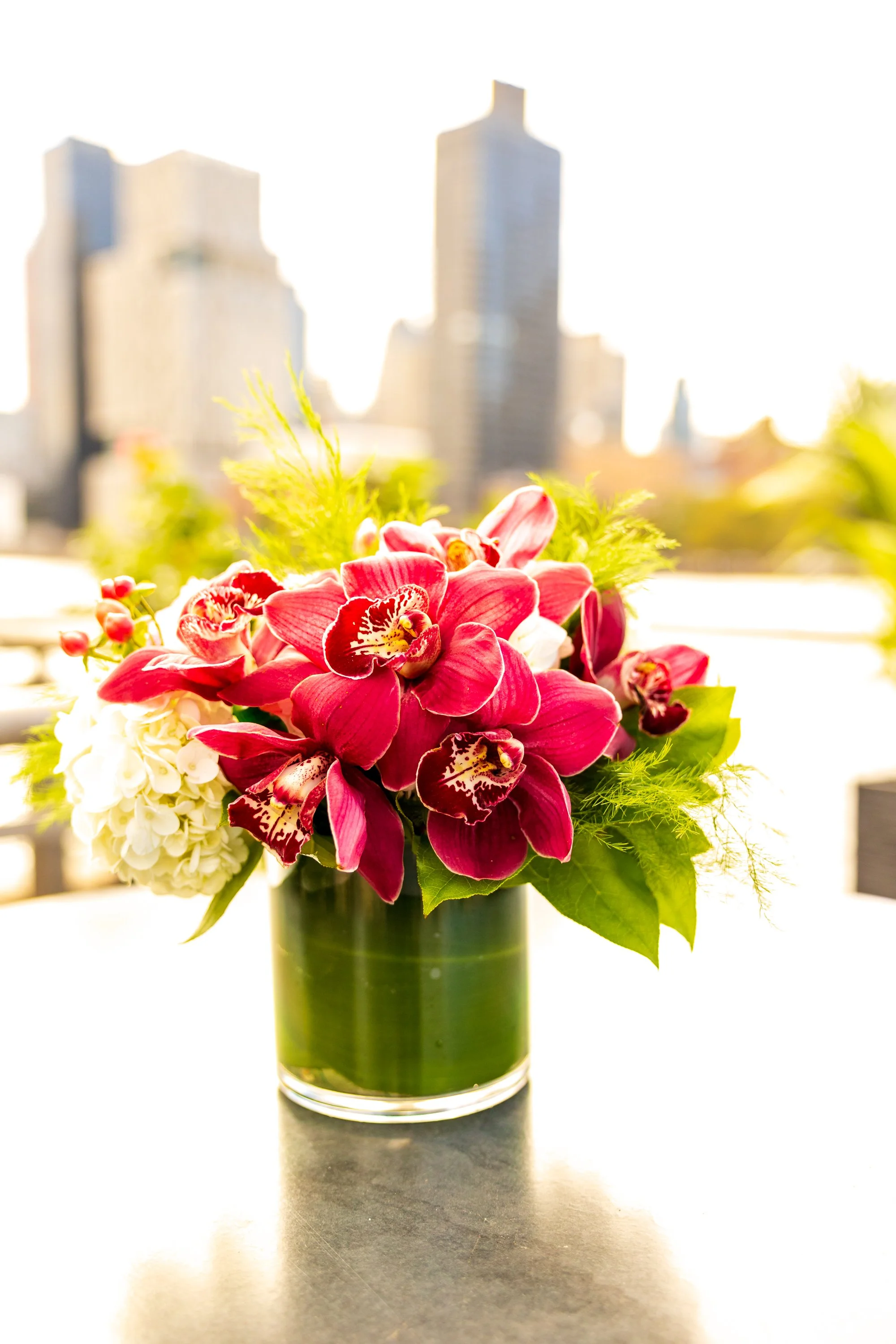 Vase of red and white orchids with green foliage on a table, blurred cityscape background.