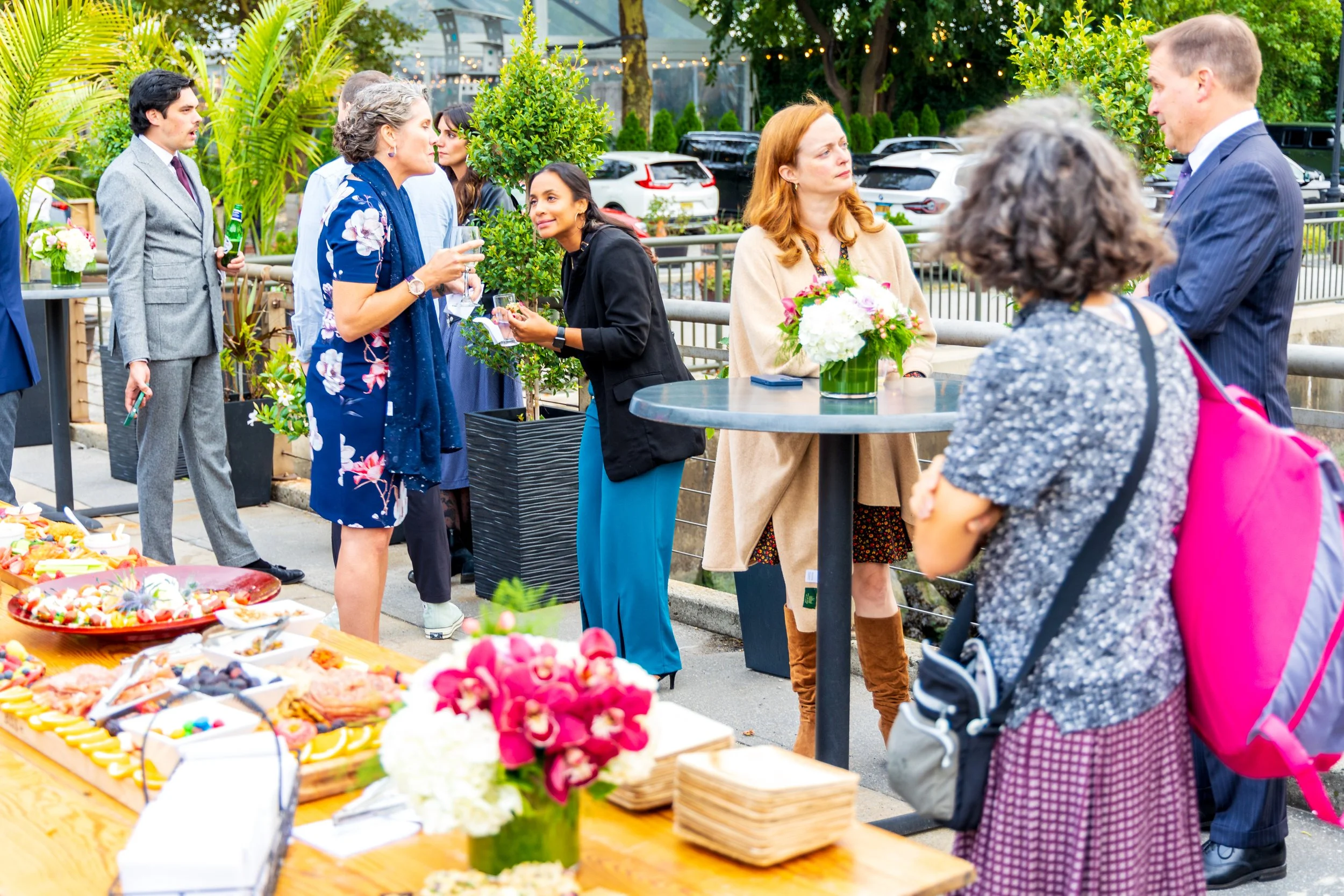 People socializing at an outdoor event with food and flowers, some holding drinks, in a green garden setting.