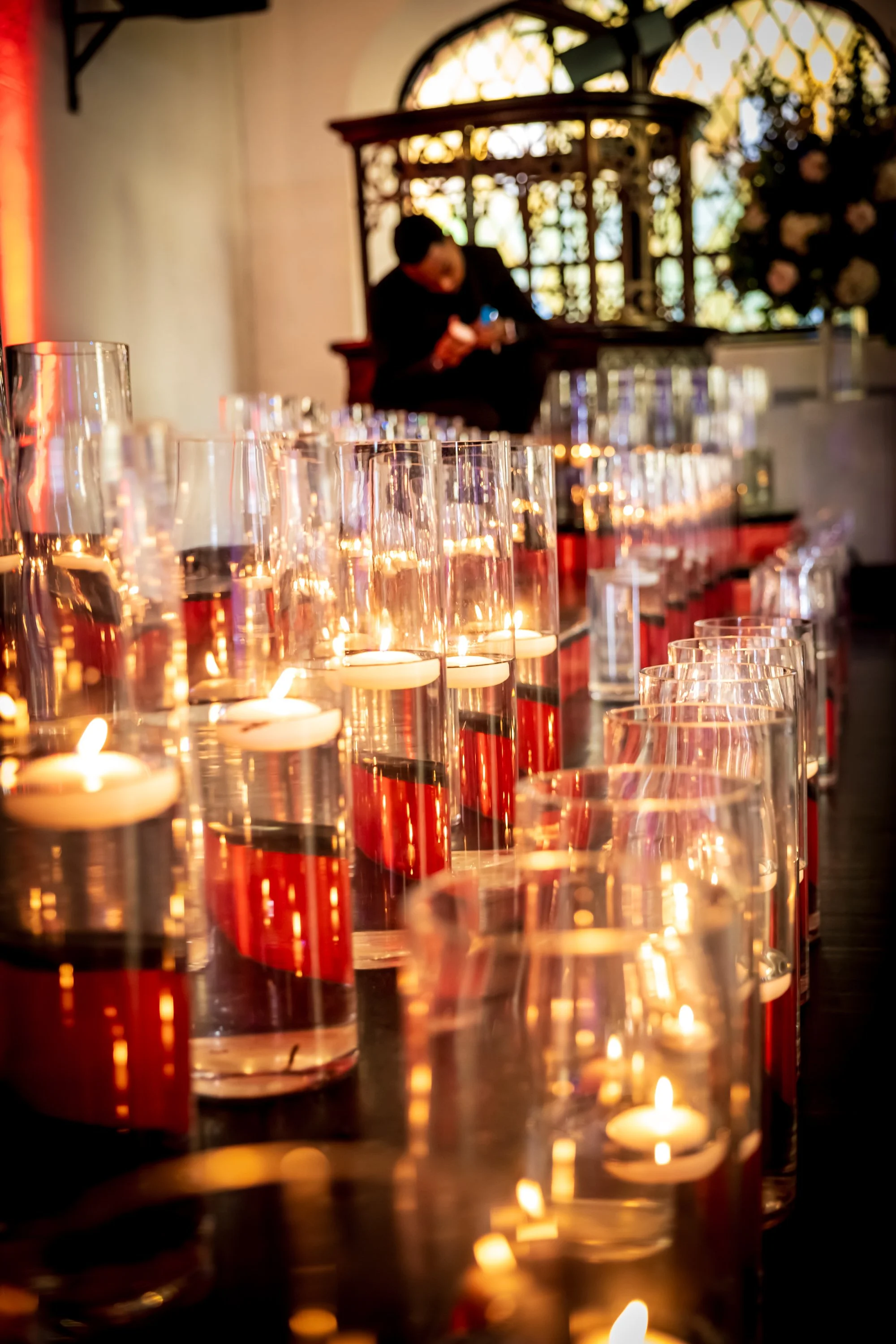 Table decorated with multiple lit candles in tall glass holders at an event, with a woman sitting in the background