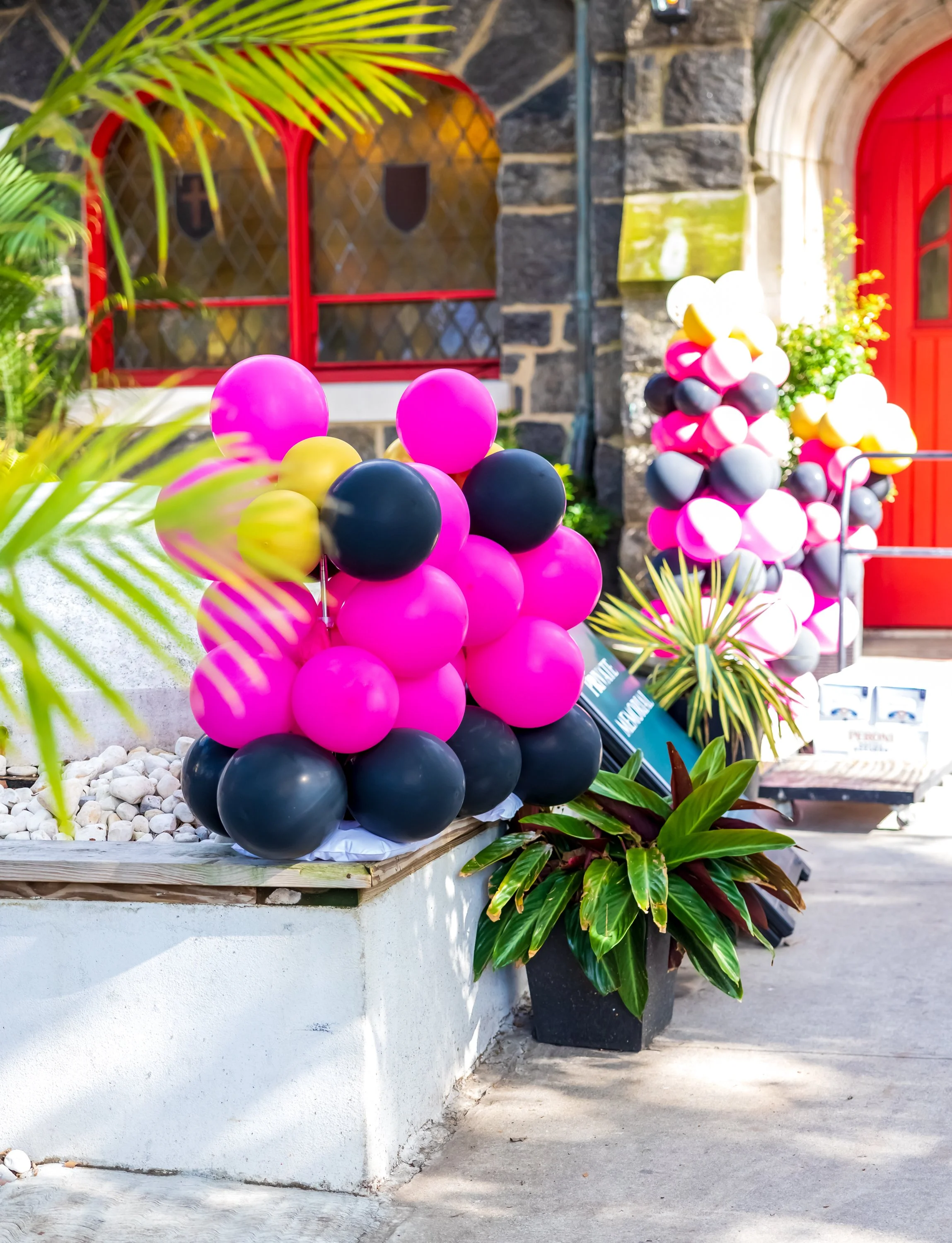 Decorative balloon arrangements in pink, black, grey, and yellow outside a building with red doors and stone walls.