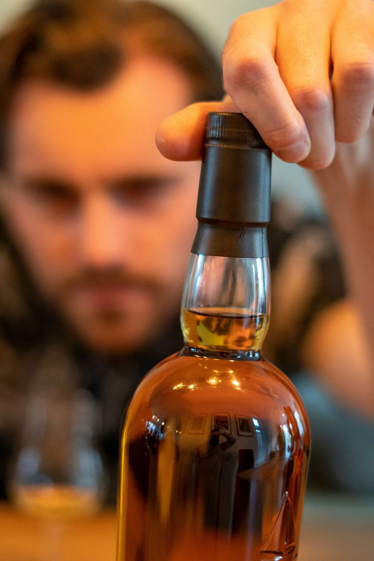 Close-up of a person's hand reaching for a whiskey bottle, with a blurred person in the background.