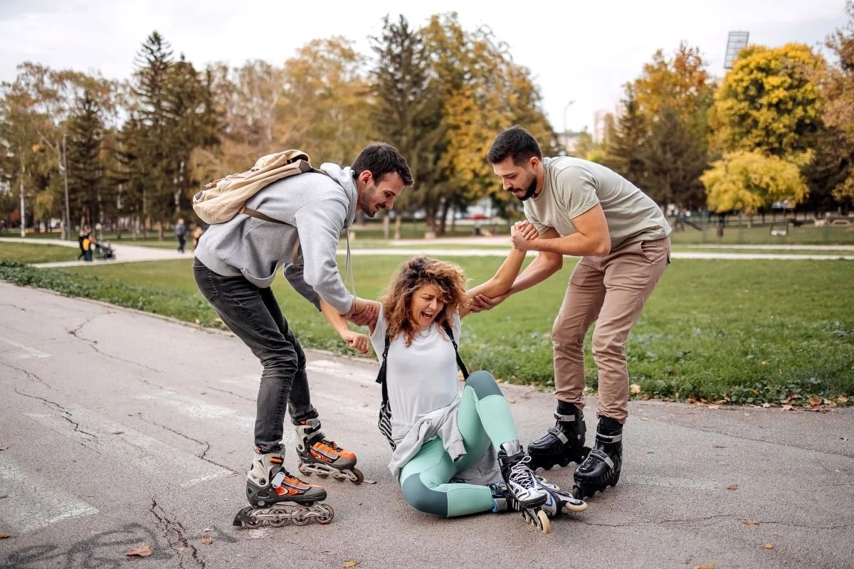 A young woman has fallen while rollerblading in a park, and two men are helping her up, holding her arms. The park has green grass and trees with autumn-colored leaves.