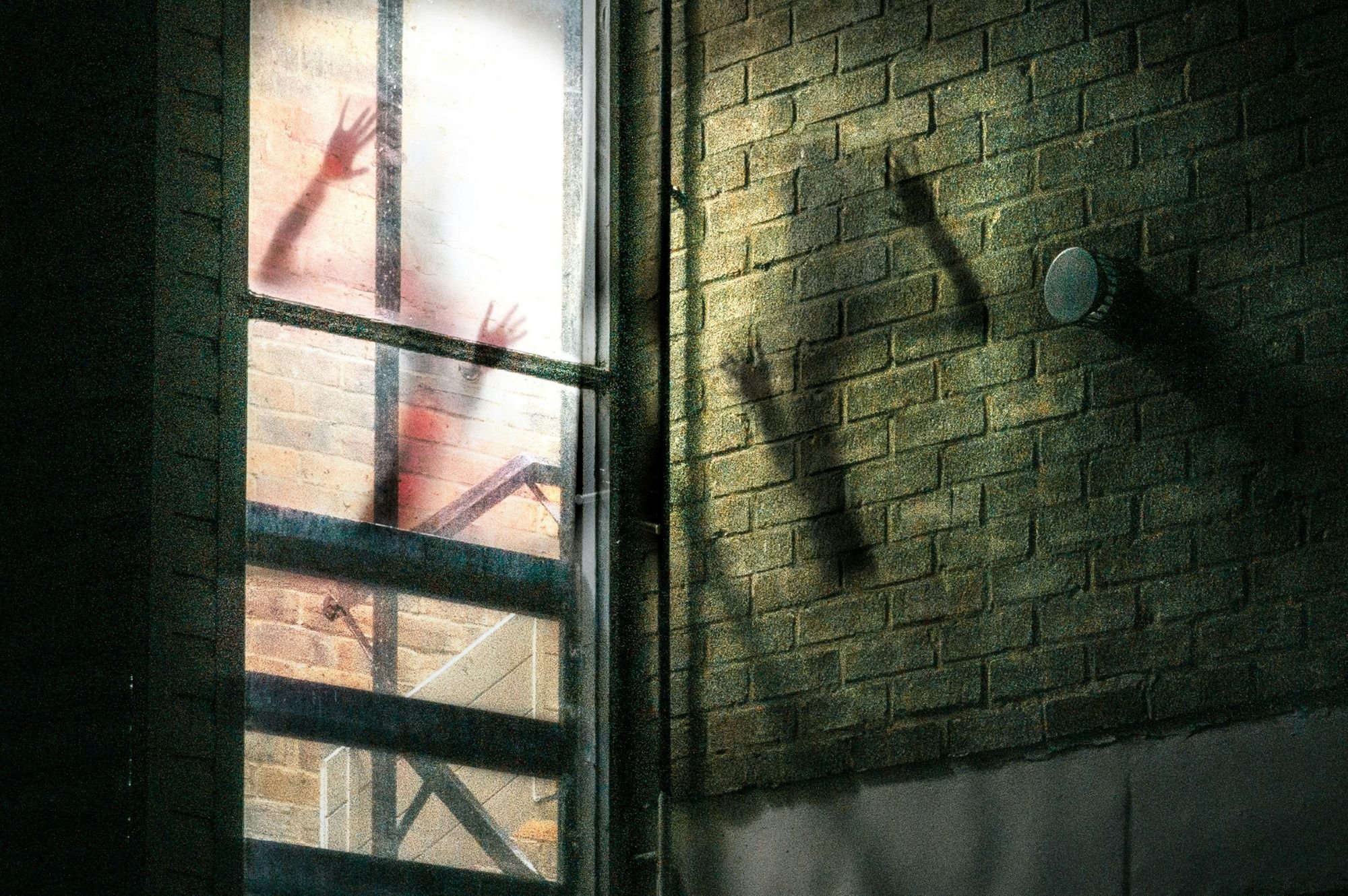 Shadow of a person with outstretched arms and hands on a foggy glass window, seen from inside, on a brick wall exterior with a vent.