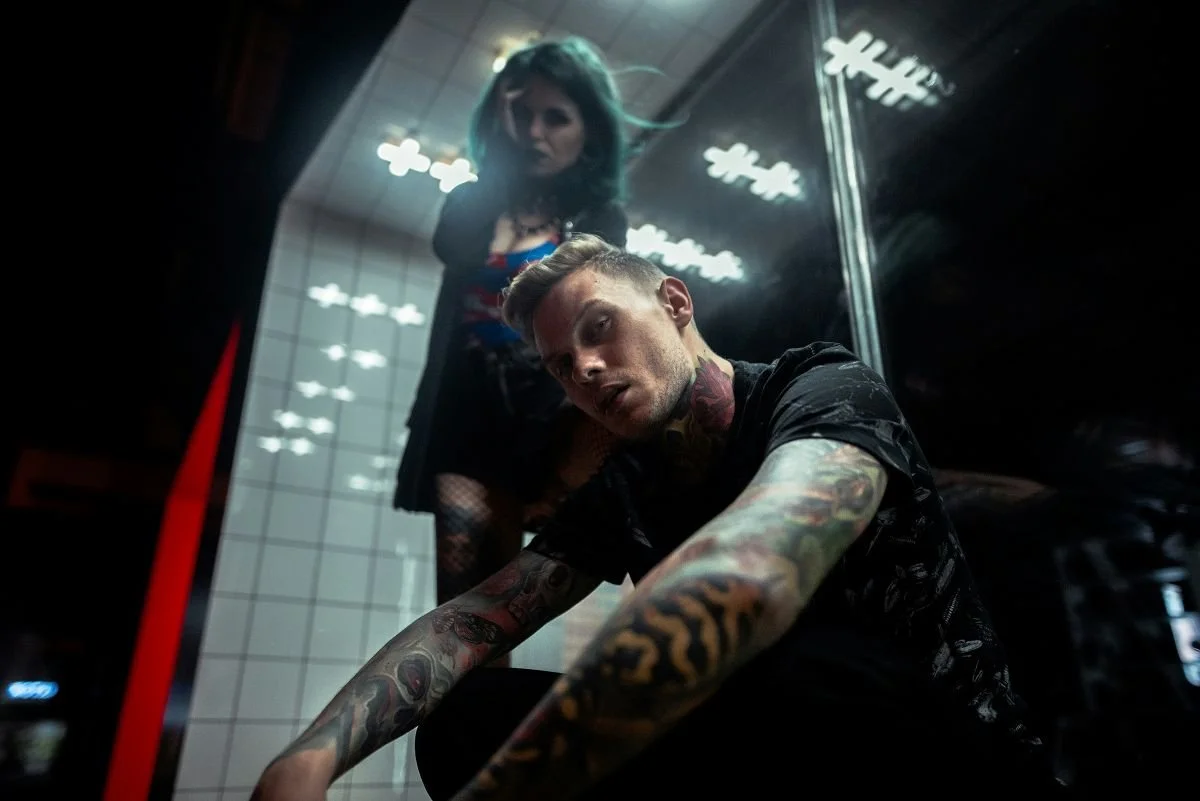 A tattooed man with short hair and an angry expression, squatting in a subway station, with a woman with blue-green hair standing behind him.