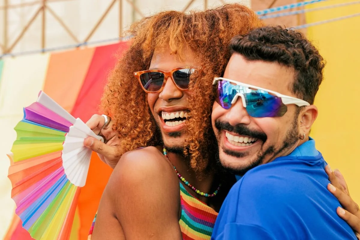 Two people in rainbow-colored clothes and sunglasses smiling and hugging at a pride event.