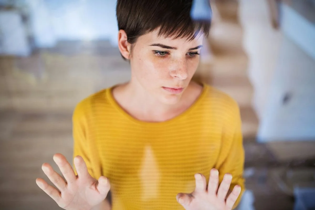 A woman with short brown hair and freckles wearing a yellow shirt, looking down with hands raised in front of her.