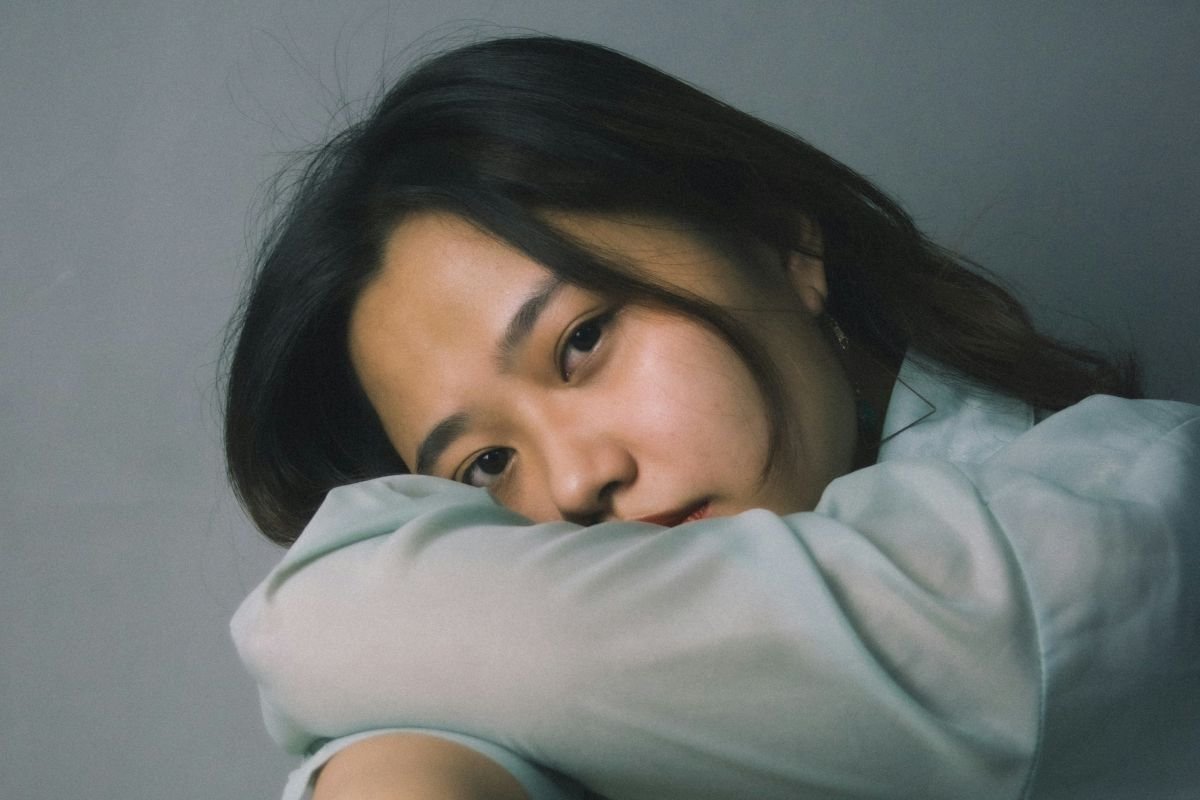 Close-up of a young woman with black hair resting her head on her arm, looking at the camera with a sad expression.