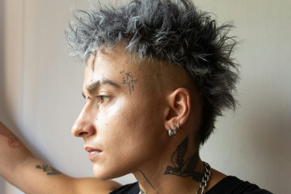 Side profile of a nonbinary person with gray and black spiked hair, multiple facial tattoos, earrings, a tattoo of a bird on their neck, and a chain necklace.
