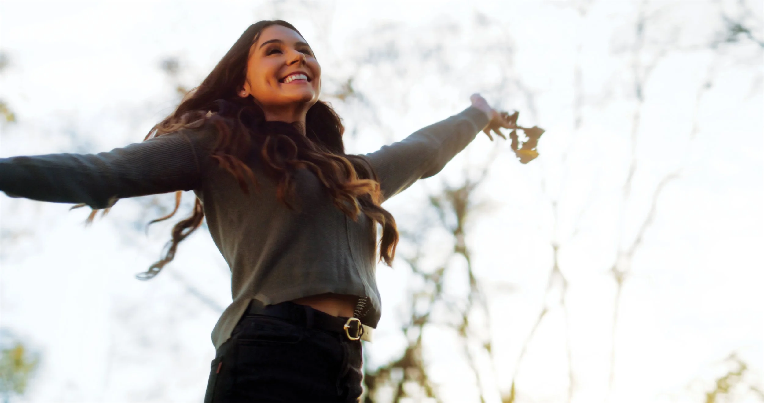 Young woman outdoors with arms outstretched, smiling, holding leaves, surrounded by trees.